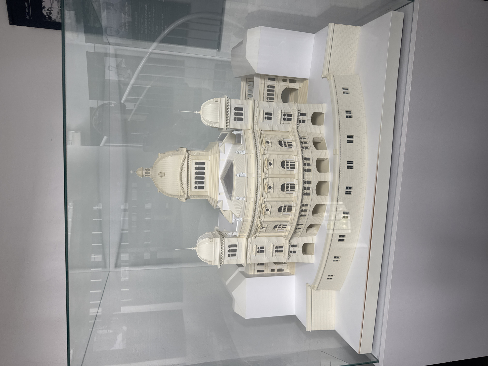 This media file shows a model of the Swiss Parliament Building in Bern, Switzerland. The model is detailed and appears to be made of a cream-colored material. It features a large central dome with a spire, smaller domes, multiple levels of windows, and arched walkways. The model is displayed inside a glass case, with reflections visible on the glass.
Behind the model, there is a black and white photograph with some text. The visible text indicates that the photo is related to "Bern boomt" (Bern is booming) and mentions "Das Parlament" (The Parliament) and "Baustelle von Westen" (Construction site from the West). Other text fragments suggest details about the building's construction period ("erbaut 1894-1902") and its location ("Standort am neu guschaffenen Bundesplatz" - Location on the newly created Federal Square).
The overall scene is that of an exhibit, likely in a museum or information center, showcasing the historical and architectural significance of the Swiss Parliament Building. The lighting is neutral, suggesting an indoor setting, possibly during the daytime. There are no people clearly visible in the main frame of the image, but the context of a historical exhibit implies a passive audience viewing the display. The model itself is the primary subject, highlighting the building's grand design.