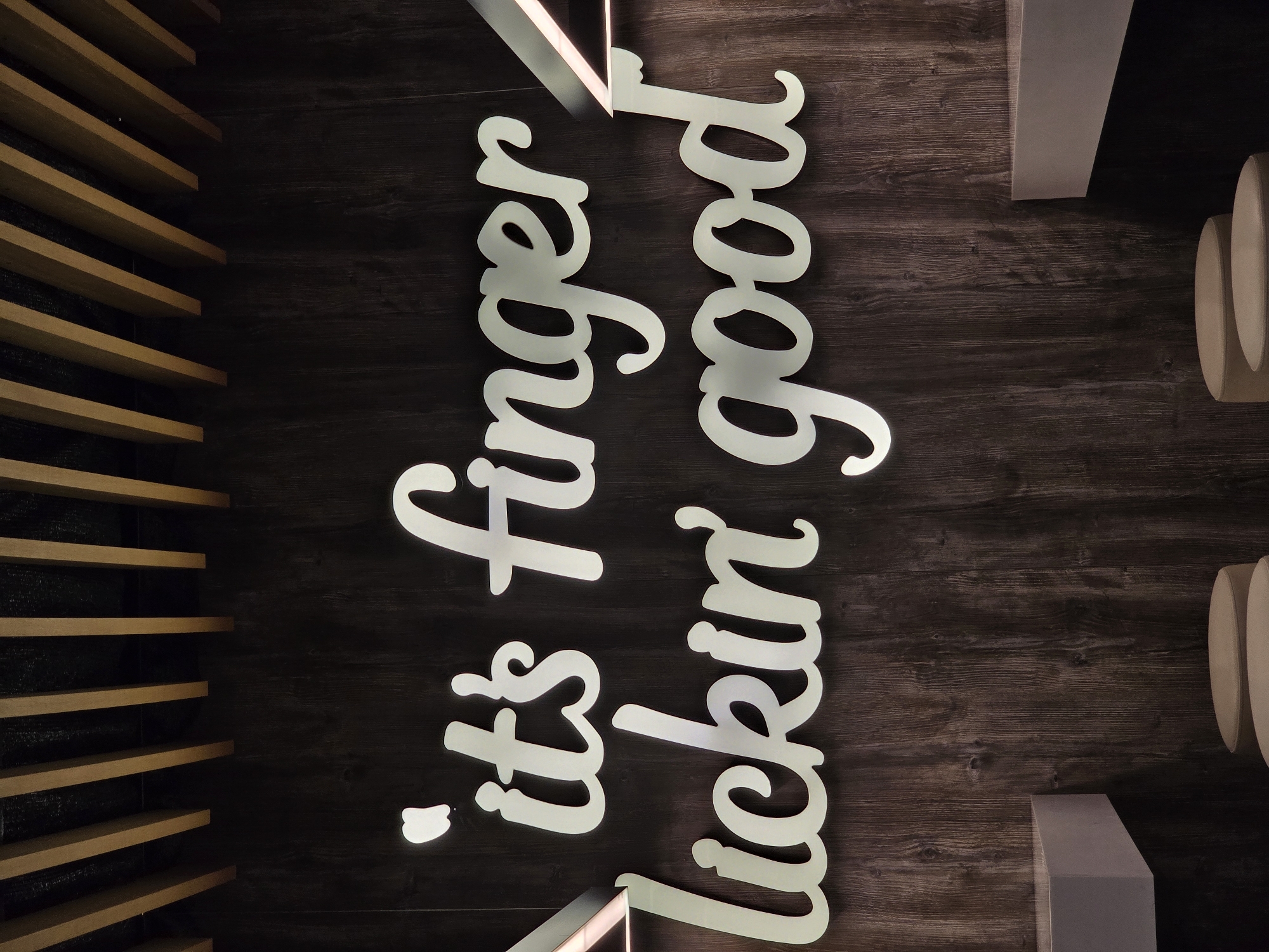 The image displays an indoor wall adorned with a large, illuminated sign, set against a dark, richly textured wooden background. The primary subject is the white or light-colored text on the sign, rendered in a flowing, cursive font, which clearly reads: "'it's finger lickin' good'". The letters appear to be dimensional or cut-out, with a soft glow around them, indicating a backlighting effect that highlights the phrase.
On the left side of the frame, a decorative architectural feature is visible, consisting of multiple horizontal wooden slats arranged in a parallel pattern, providing a contrasting texture to the dark wall. To the right, partially seen, are light-colored, cylindrical elements stacked vertically, which could be part of a decorative column, a lamp, or another piece of interior furnishing. Below these, a light-colored, solid rectangular block is also visible.
The overall ambiance of the scene is dim, suggesting an intimate or cozy setting, often characteristic of a restaurant or cafe interior. The lighting primarily draws attention to the sign. There are no people present, and no specific activity is occurring beyond the display of the sign itself. The phrase on the sign is famously associated with the fast-food chain KFC, strongly indicating that the location is a branch of this restaurant in Sofia, Bulgaria, as per the provided context. The image focuses on the interior design and branding elements.