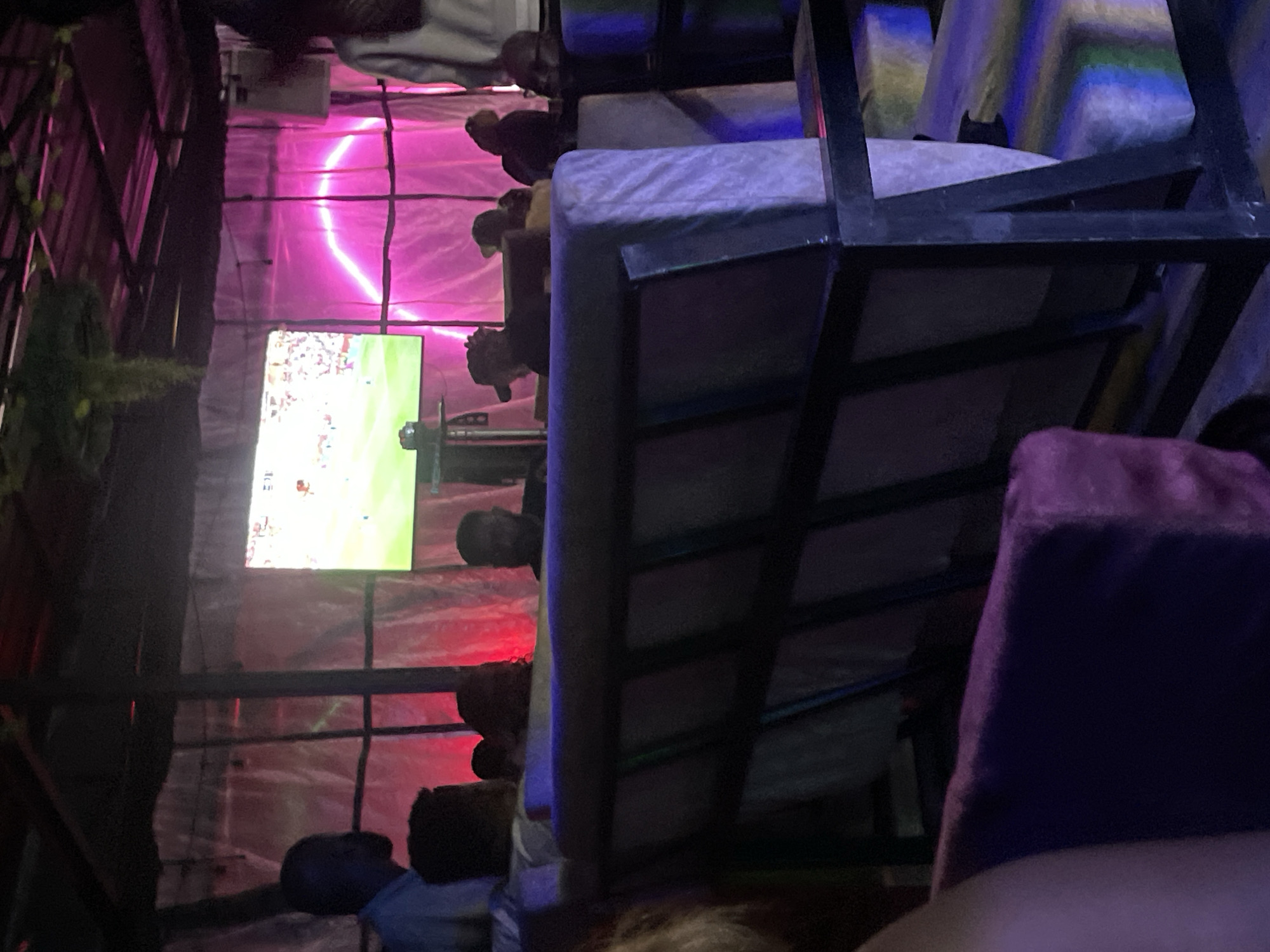 The image depicts an indoor scene, likely a lounge or bar, with a large television screen displaying a sporting event. The room is dimly lit with ambient pink and purple lighting, creating a vibrant and somewhat mysterious atmosphere. Several people are visible, seated and standing, some appearing to be watching the screen. The furniture includes couches and what appear to be cushions, some with colorful striped patterns. The background features translucent white or clear sheeting, possibly part of the room's decor or enclosure, with a prominent neon pink curved light fixture. The overall impression is a social gathering space, potentially a viewing lounge for sports enthusiasts, located in Gura Top, Nigeria.