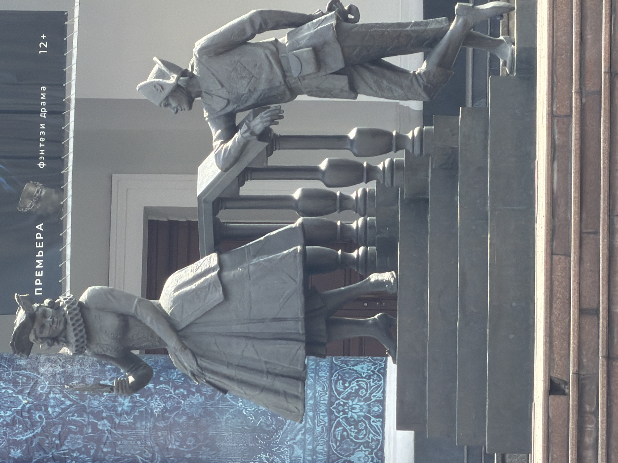 The image depicts a monument in Krasnoyarsk, Russia, featuring two stylized bronze statues of characters from Nikolai Gogol's play "The Inspector General." One statue, representing Khlestakov, leans over a stylized balustrade and staircase. The other, representing Anna Andreyevna, stands below, in front of a poster with Cyrillic text advertising a theatrical performance. The backdrop includes a light-colored building facade and a section of a fluted column. The statues are rendered in a simplified, almost cartoonish style.