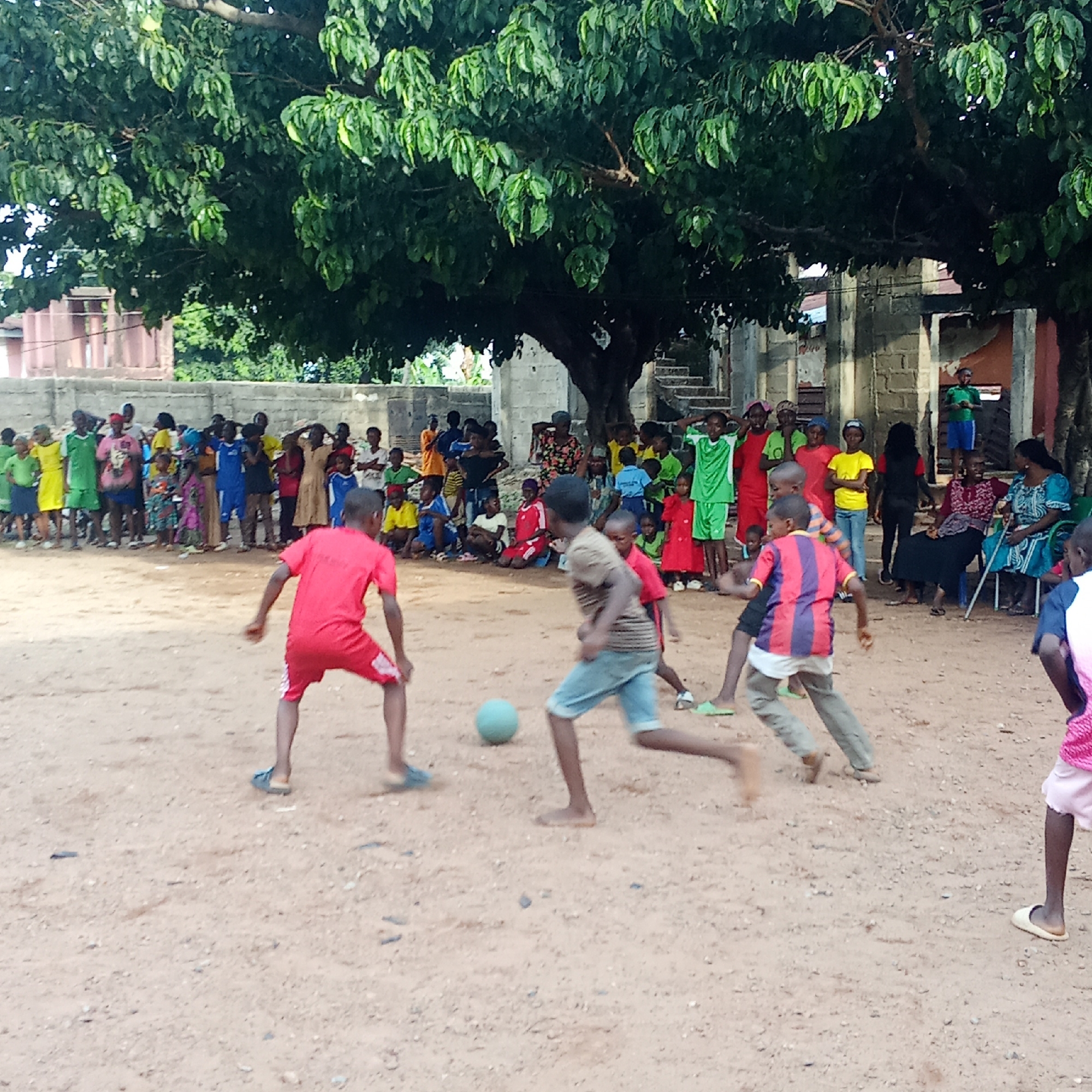 The image depicts a group of children playing soccer on a dirt field in Akwanga, Nigeria. Several adults are seated and standing around the perimeter of the field, observing the game. The players are dressed in casual clothing. A concrete building stands in the background, partially obscured by a large tree. The ground is sandy and uneven.