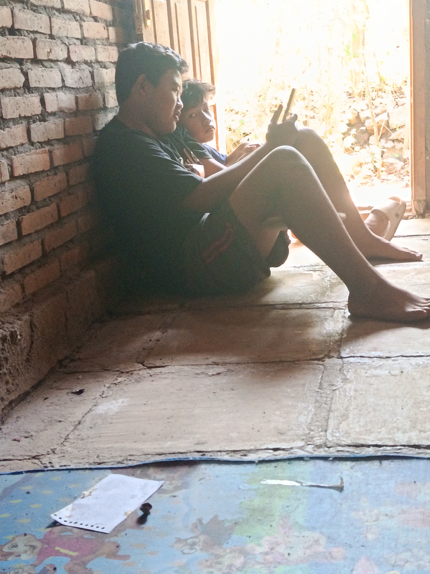 Two young individuals, likely adolescent males, are seated against a brick wall. The individual on the left is wearing a dark t-shirt and appears to be looking towards the right. The individual on the right is leaning into the person on the left, with their face partially obscured. They are both barefoot, and the person on the right is wearing a slipper on their left foot. The person on the left is holding a mobile phone. The environment suggests an outdoor or semi-outdoor setting, with sunlight streaming in from an open doorway or window, illuminating foliage and debris outside. The floor is tiled. In the foreground, a crumpled piece of paper with perforated edges and some dark smudges is visible on a colorful mat. A bent nail is also present on the mat. The scene is located in Karangmojo, Indonesia.