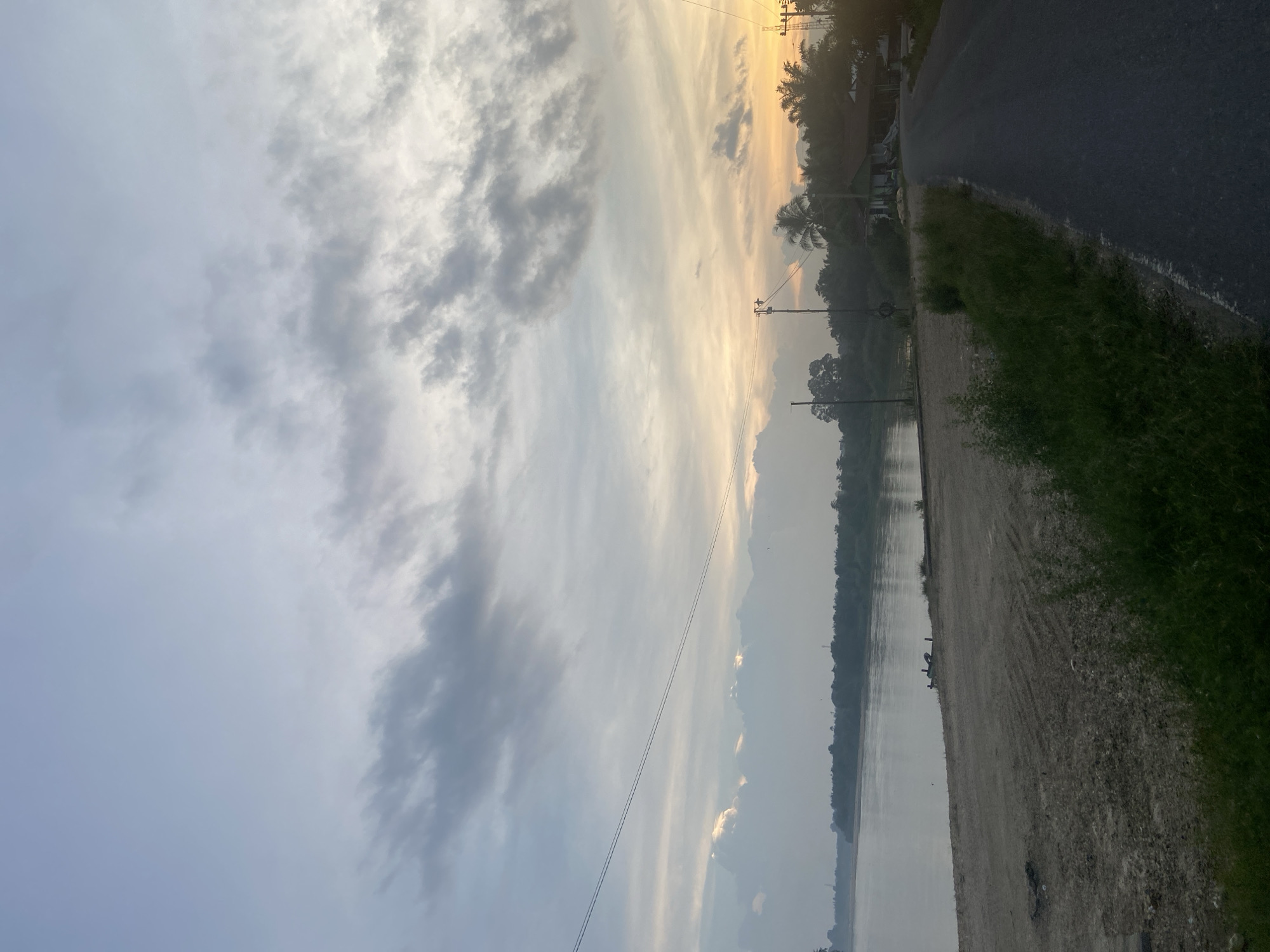 The image depicts a scenic view of a road alongside a body of water, possibly a river or lake, in Lembah Dusun Gading, Indonesia. The scene is bathed in the soft, warm light of either sunrise or sunset, indicated by the golden hues in the sky and the elongated shadows. Wispy clouds stretch across the sky, adding texture and depth to the atmosphere.
On the right side of the frame, an asphalt road runs alongside a strip of green grass. Beyond the grass, the water stretches out, reflecting the muted colors of the sky. The left bank of the water is lined with lush green vegetation and trees, creating a natural boundary. In the distance, a faint silhouette of mountains can be seen through a slight haze, suggesting a tranquil, natural setting.
Several utility poles with power lines are visible along the roadside and extending across the landscape, hinting at human presence and infrastructure in this otherwise natural environment. The overall mood of the image is peaceful and serene, capturing the quiet beauty of the Indonesian landscape at a transitional time of day. No people are clearly discernible in the image, but a couple of figures appear to be near the water's edge on the sandy bank. The image is oriented vertically, so the scene is viewed as if the camera was tilted.