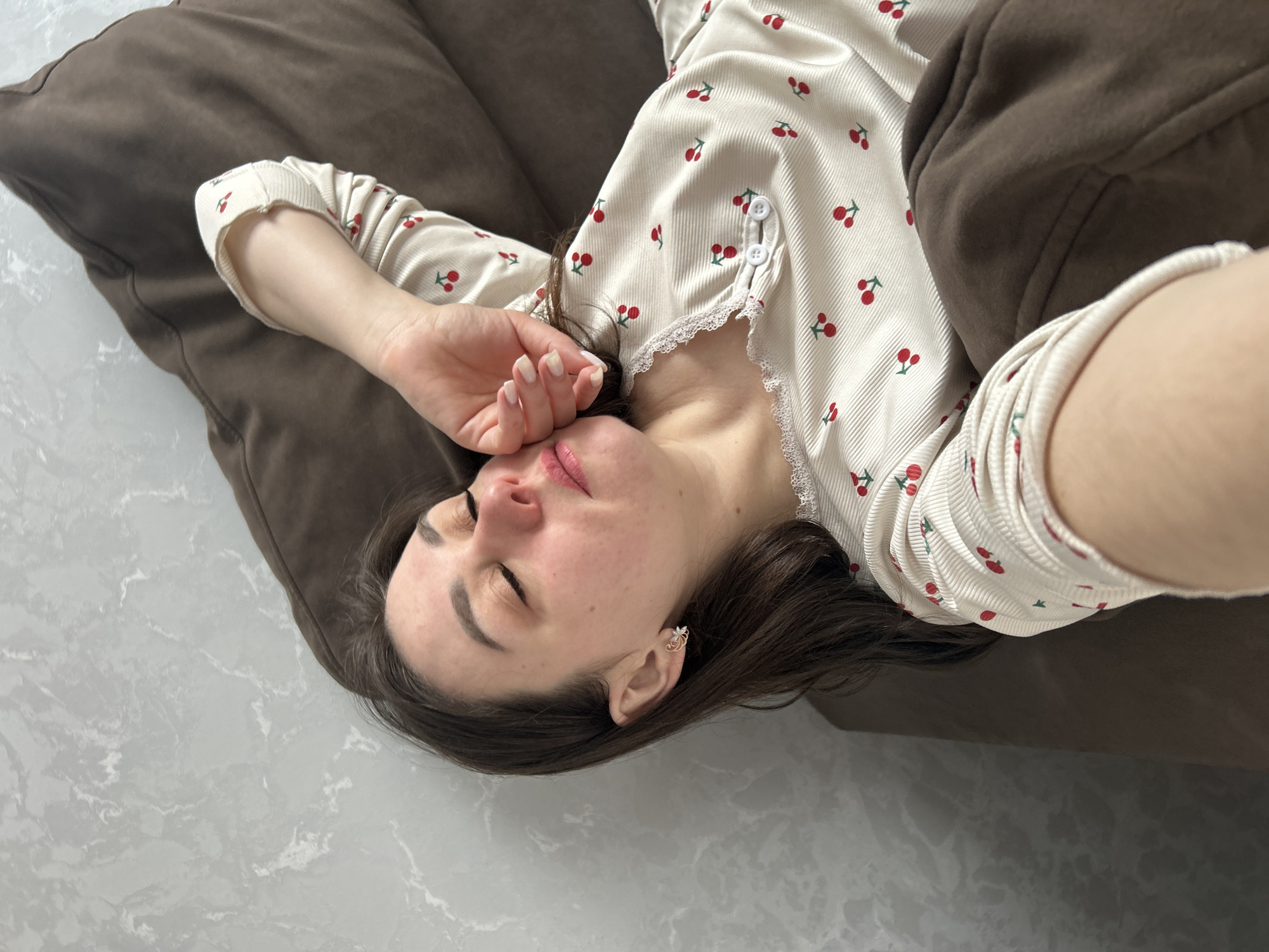 A young woman lies on a brown pillow on a gray marble floor. She is wearing a white pajama top with a cherry print and lace trim. Her eyes are closed, and her hand is near her face. She has dark hair and is wearing an earring. The location is Chaikovsky, Russia.