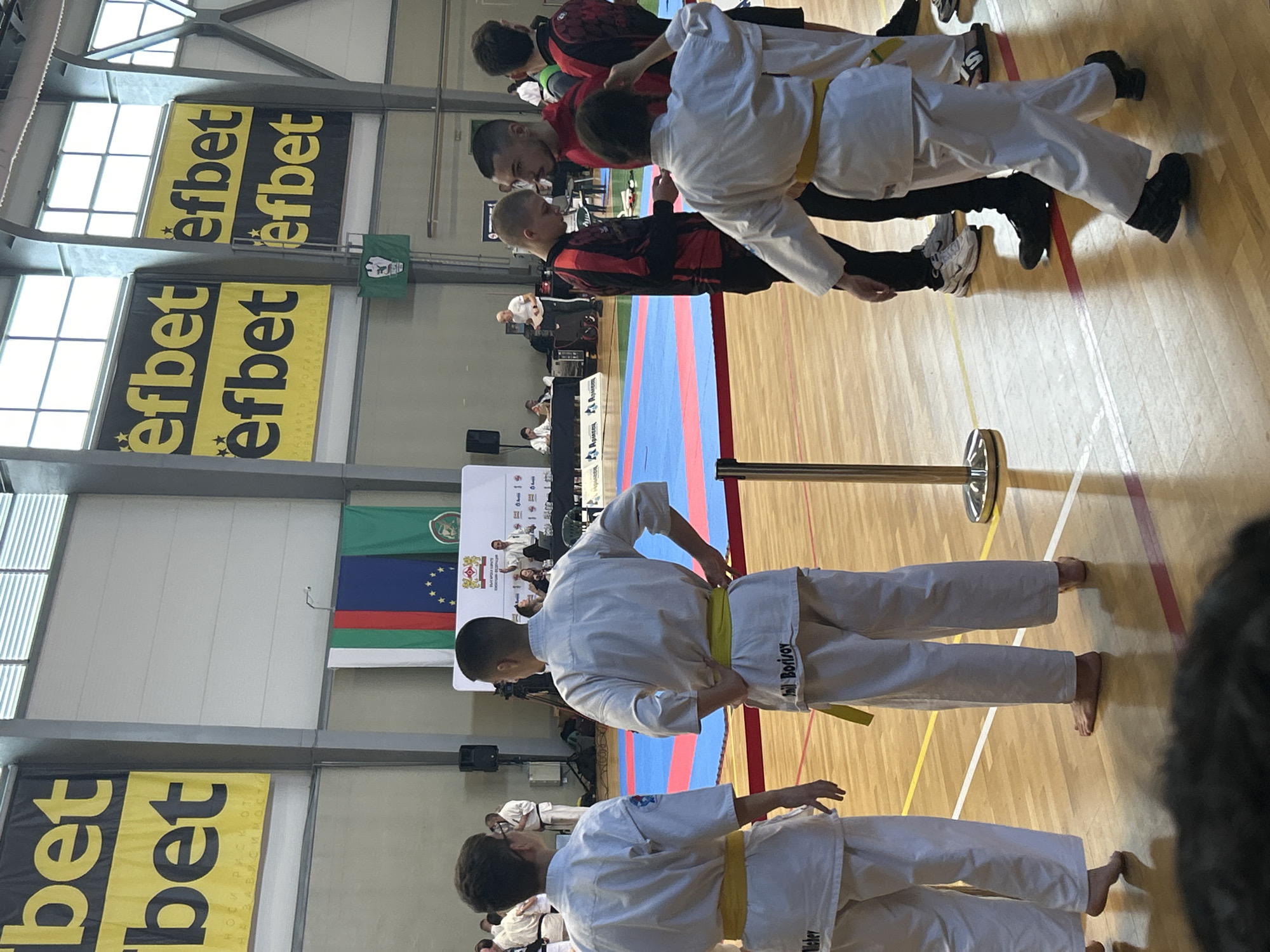 The image depicts an indoor martial arts competition, likely karate or similar discipline. Several participants are seen, some in white uniforms with yellow belts and others in red and black attire. The competition appears to be taking place on a wooden floor marked with lines, and a blue and red mat is in the background. Banners with the text "efbet" are visible along the walls. Flags of Bulgaria and the European Union, along with other flags, are displayed near the center of the room. The text "Borisov" and "Weber" are visible on the uniforms of some participants. Based on context clues, the event may be held in Stara Zagora, Bulgaria.