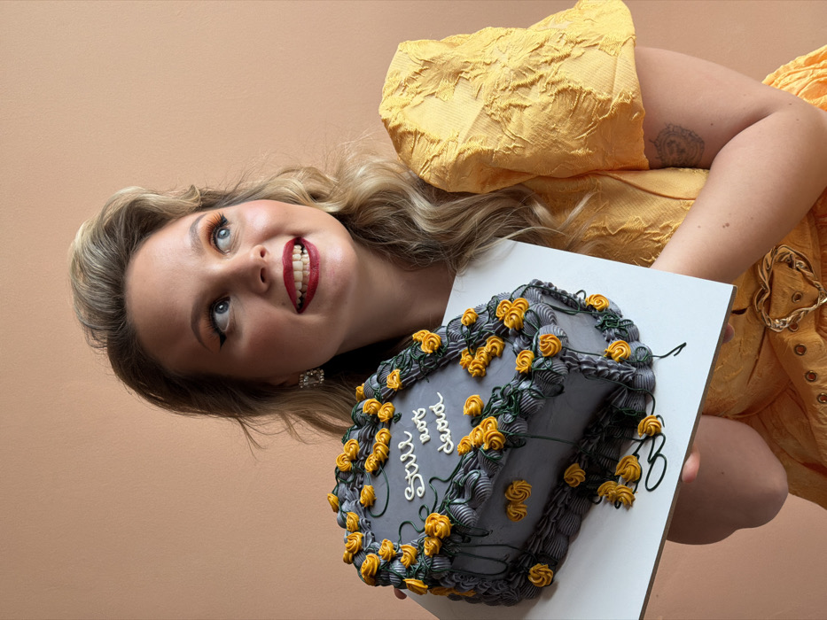 A blonde woman, appearing to be in her late twenties to early thirties, is positioned from the mid-torso up against a plain light beige background. She has styled hair, a full face of makeup including orange eyeshadow, black winged eyeliner, and red lipstick, and wears a decorative earring on her visible left ear. She is dressed in a yellow, textured dress with puffy short sleeves and a gold heart-shaped buckle at the waist. A faint tattoo is visible on her right upper arm. She is holding a dark grey, coffin-shaped cake decorated with numerous small yellow icing roses and green icing vines. The cake has white icing text on its top surface that reads "Still not dead" and rests on a white rectangular board. The woman is smiling broadly, with her mouth slightly open, and her gaze is directed upwards and to the right, away from the cake. The background provides no discernible details to indicate a specific location within Tel-Aviv, Israel.