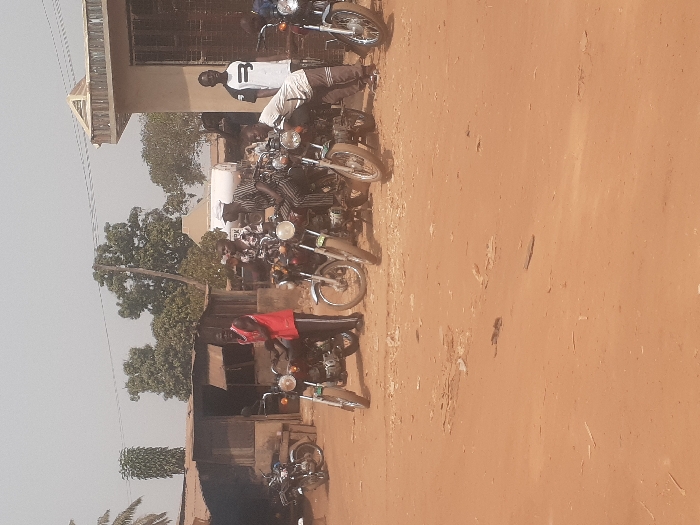 This outdoor scene is located in the city of Gwadanye, Nigeria, featuring a dusty, reddish-brown dirt ground. Six adult males are positioned around five parked motorcycles in the mid-ground. From left to right: a male in a red sleeveless shirt and dark pants stands leaning against a dark-colored motorcycle; next, a male in a patterned short-sleeve shirt and a light cap is seated on another motorcycle; adjacent to him, a male in a striped long-sleeve shirt and a dark cap is seated on a third motorcycle. Further right, a male in a white t-shirt with a dark graphic stands near a fourth motorcycle. To his right, a male in a light t-shirt and dark pants is bent over a fifth motorcycle, appearing to service it. A sixth male is partially visible behind the rightmost motorcycle. In the background, a rustic structure with corrugated metal roofing is on the left, and a light-colored building with a dark window and decorative roofline is on the right. Overhead power lines, scattered trees, and a white cylindrical tank marked with letters like "K PK" are also visible. A sixth motorcycle is partially seen parked on the far left, near the rustic structure.