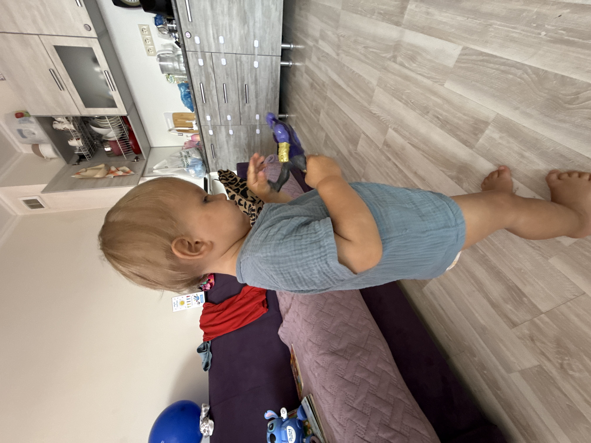 A young child, appearing to be around two years old, stands on a light-colored laminate floor in what appears to be a domestic setting. The child has fair hair and is wearing a light blue, short-sleeved, textured shirt and a diaper. They are holding a dark purple toy, possibly a water gun, with gold accents in their hands, looking down at it.
In the background, a section of a living area is visible. To the left, a dark purple sofa is present, with a blue balloon resting on it. A red garment and a blue shirt are also draped over the sofa. Further to the left, a small blue toy figure is on a book. Behind the sofa, a wall is painted a light neutral color.
The right side of the image reveals a kitchen area with light wood-toned cabinets and drawers. A white countertop is visible, with various kitchen items, including a kettle, bottles, and what appears to be a sink with a faucet. The kitchen cabinetry extends across the width of the visible space. The overall lighting suggests it is daytime. There are no visual cues to specifically place this scene in the city of Sochi, Russia.