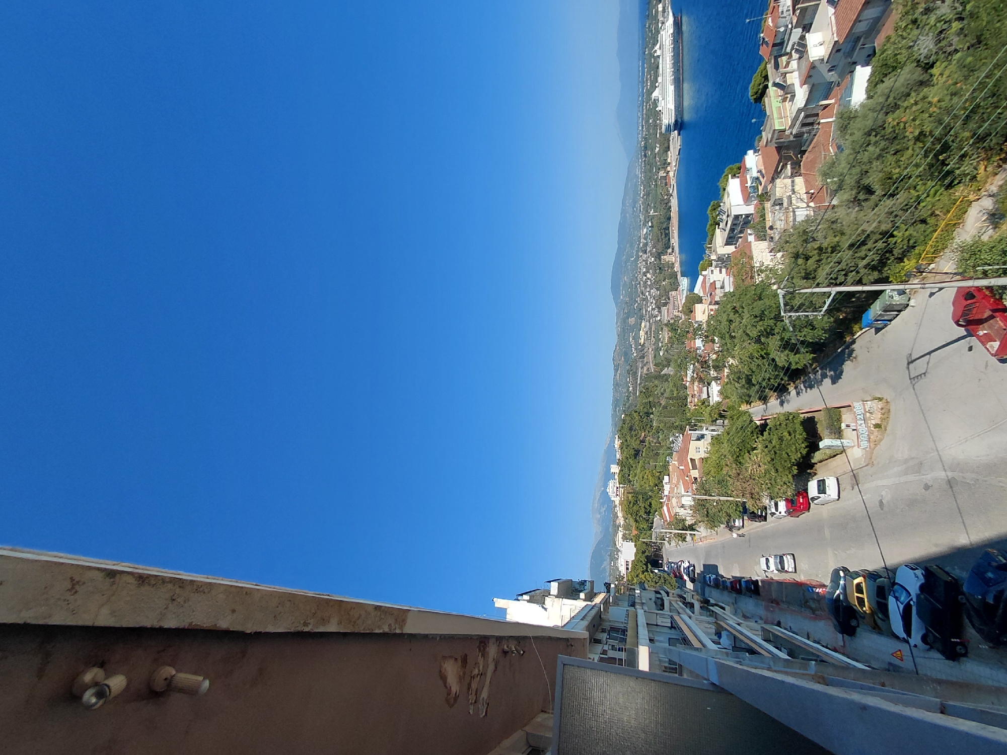 The image depicts a daytime scene overlooking a coastal city, likely in the Municipal Unit of Aegio, Greece, given the presence of a large cruise ship in the harbor and the architectural style of the buildings. In the foreground, a section of a building's balcony is visible on the left, with exposed concrete and some peeling paint. Below the balcony, a street runs downhill, lined with parked cars. Further down the street, houses with red-tiled roofs and greenery are scattered across the landscape, leading towards the blue sea. The sea is calm, with a large white cruise ship docked or sailing in the distance. The sky is a clear, vibrant blue. The overall impression is of a sunny, Mediterranean town.