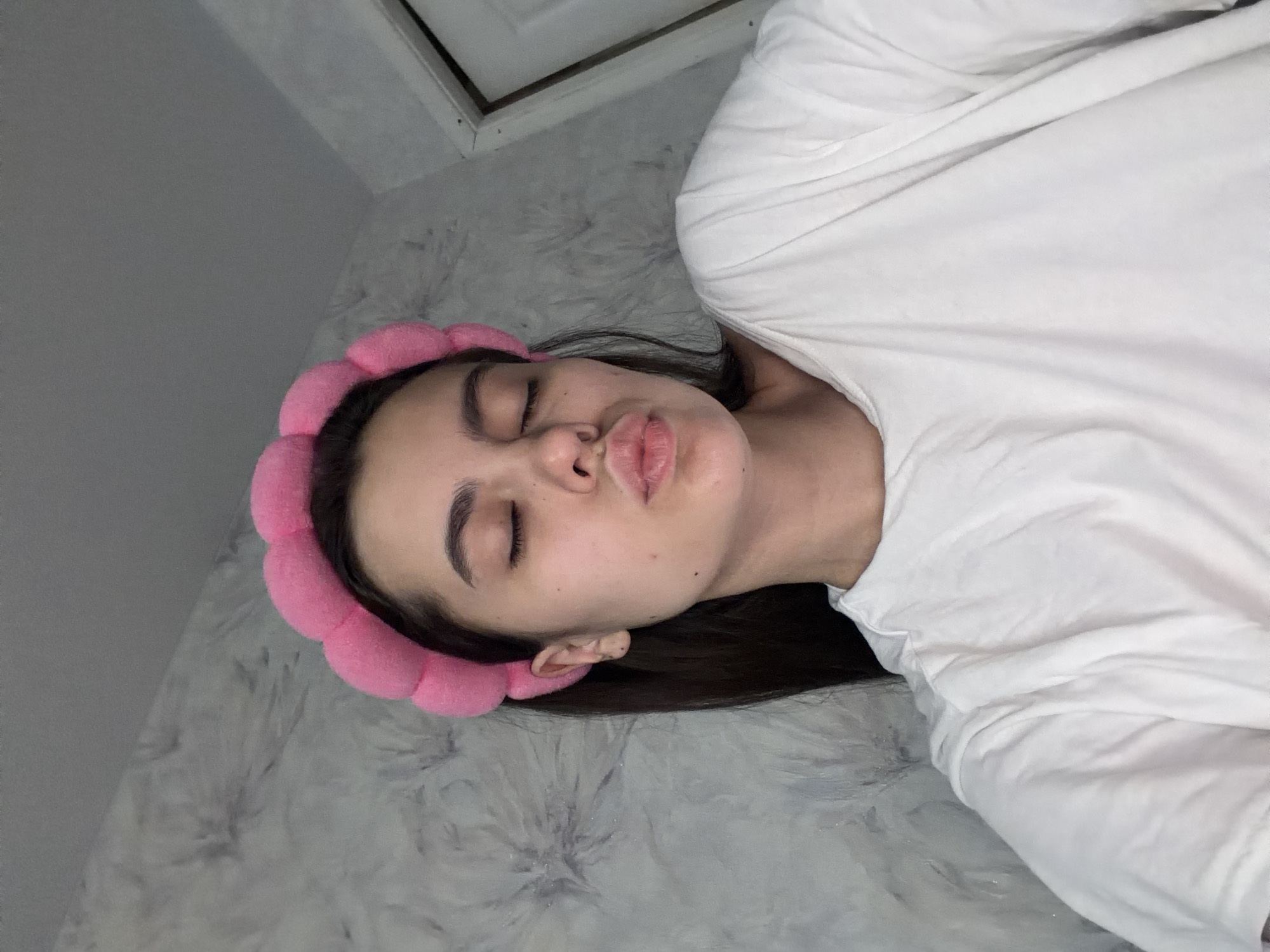 A young woman with brown hair is lying down. She is wearing a white t-shirt and a pink headband. Her eyes are closed, and she has a slight pout on her lips. The background appears to be a patterned wall, possibly in a bathroom. The location is unknown and the city of Chaikovsky, Russia, is not confirmed.