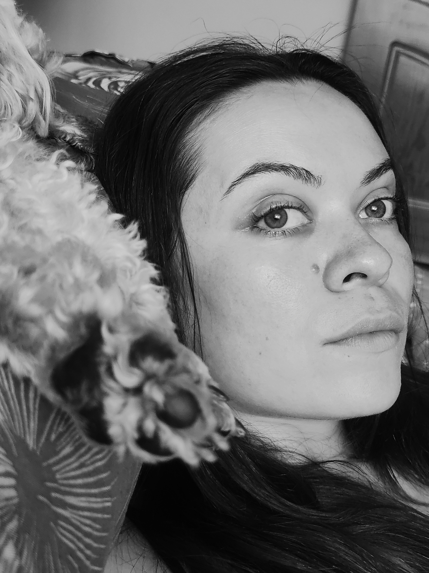 A black and white close-up portrait shows a woman with dark hair and light skin looking upward with a serious expression. The woman has a mole on the right side of her nose. A dog's paw with black pads is resting on her left shoulder. The background includes a patterned fabric. The photo was taken in Chaikovsky, Russia.