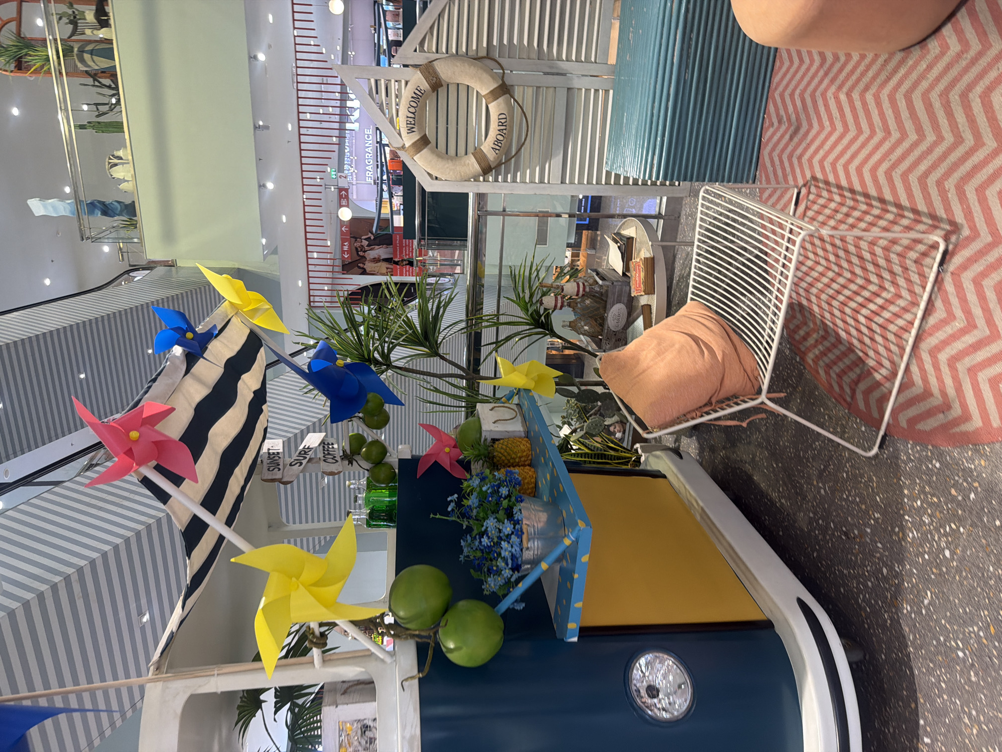 In the city of Wichit, Thailand, a brightly decorated indoor scene resembles a beachside refreshment stand. A dark blue and yellow van, reminiscent of a classic camper, serves as the focal point. Brightly colored pinwheels in pink, yellow, and blue spin in the air. Green spherical fruits hang from a branch, and a bucket of blue flowers sits on a small shelf, accompanied by pineapples and a white paper bag. A white metal chair with a peach-colored cushion is placed on a rug with a pink and white chevron pattern. Behind the chair, a white shutter-style panel displays a life preserver with the word "WELCOME" and "ABOARD". A teal corrugated structure is positioned to the right of the chair. The background suggests a retail environment, with railings, signage, and various displays visible on a higher level, indicating this is likely a promotional display within a shopping mall or similar establishment.