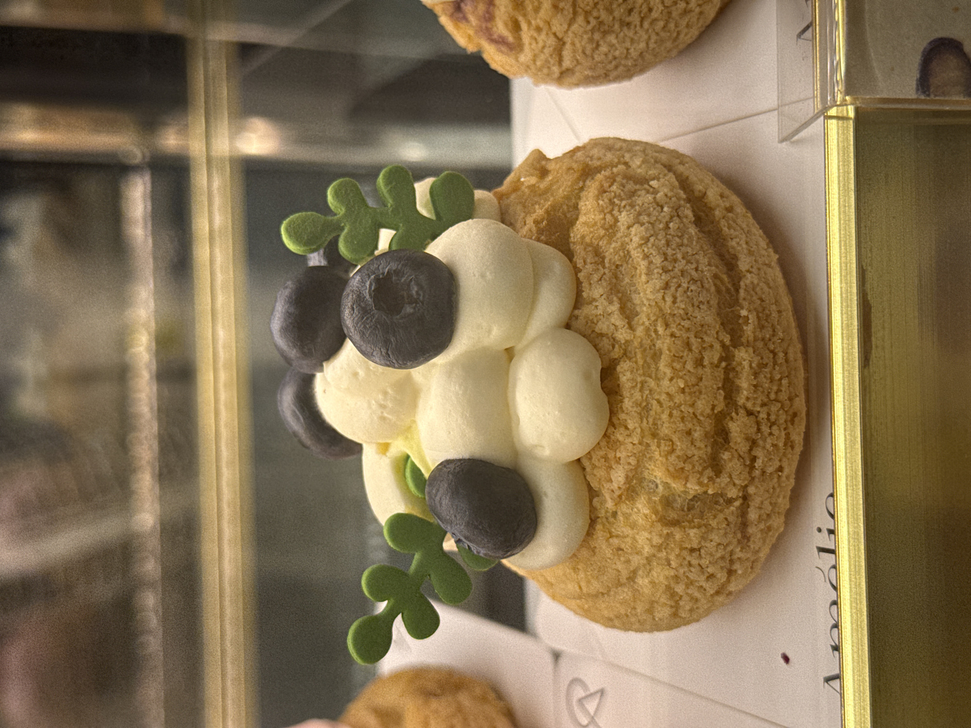 An eye-level, indoor shot shows a cream puff pastry with blueberries and green decorations on top, sitting on a box with the name "Amélie" printed on it in Da Lat, Vietnam. The cream puff has a rough, golden-brown surface. White piped cream forms a base for three dark blueberries and two green, decorative leaves. The pastry sits on a white box with faint markings and the name "Amélie" printed vertically in dark ink. A gold-colored metal frame surrounds the box. A glass display case is visible in the background, reflecting the surrounding environment. Another pastry sits out of focus at the top of the frame, and a portion of another pastry is also visible in the lower part of the frame.