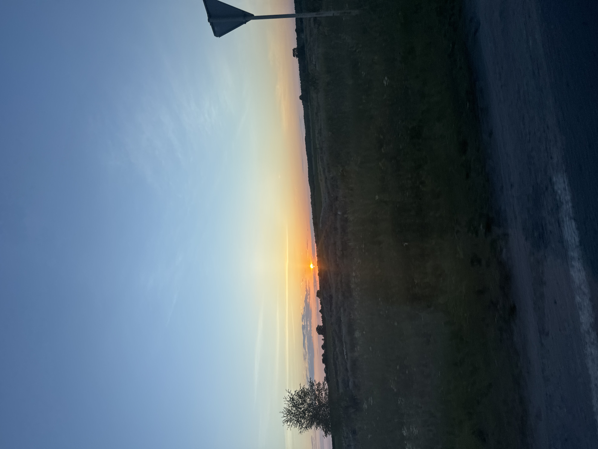 The image depicts a rural landscape in Orlivka, Ukraine, during sunset. The sky transitions from a clear light blue at the top to warm orange and pink hues near the horizon. A solitary tree stands on the left side of the frame, silhouetted against the colorful sky. The middle ground is a field of tall grass and scattered bushes. A road with a white line is visible in the foreground. On the right side of the image, a triangular road sign on a pole is partially visible against the sky. The overall scene conveys a sense of tranquility and natural beauty as day transitions into night.