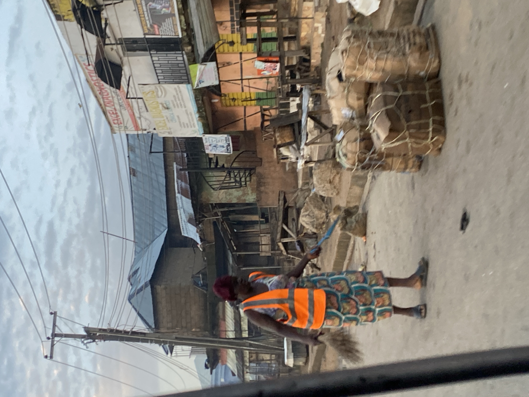 A woman wearing an orange high-visibility vest over a patterned dress sweeps the street with a broom. She is on a paved street in a commercial area. Behind her, buildings with corrugated metal roofs are visible. Signs for "EXCEL DIAGNOSTICS CLINICAL SERVICES" are displayed on one of the buildings. Several large bundles wrapped in netting are stacked on the side of the street. Utility poles with overhead wires are also present. The sky is overcast. This scene is located in the city of Gwong, Nigeria.