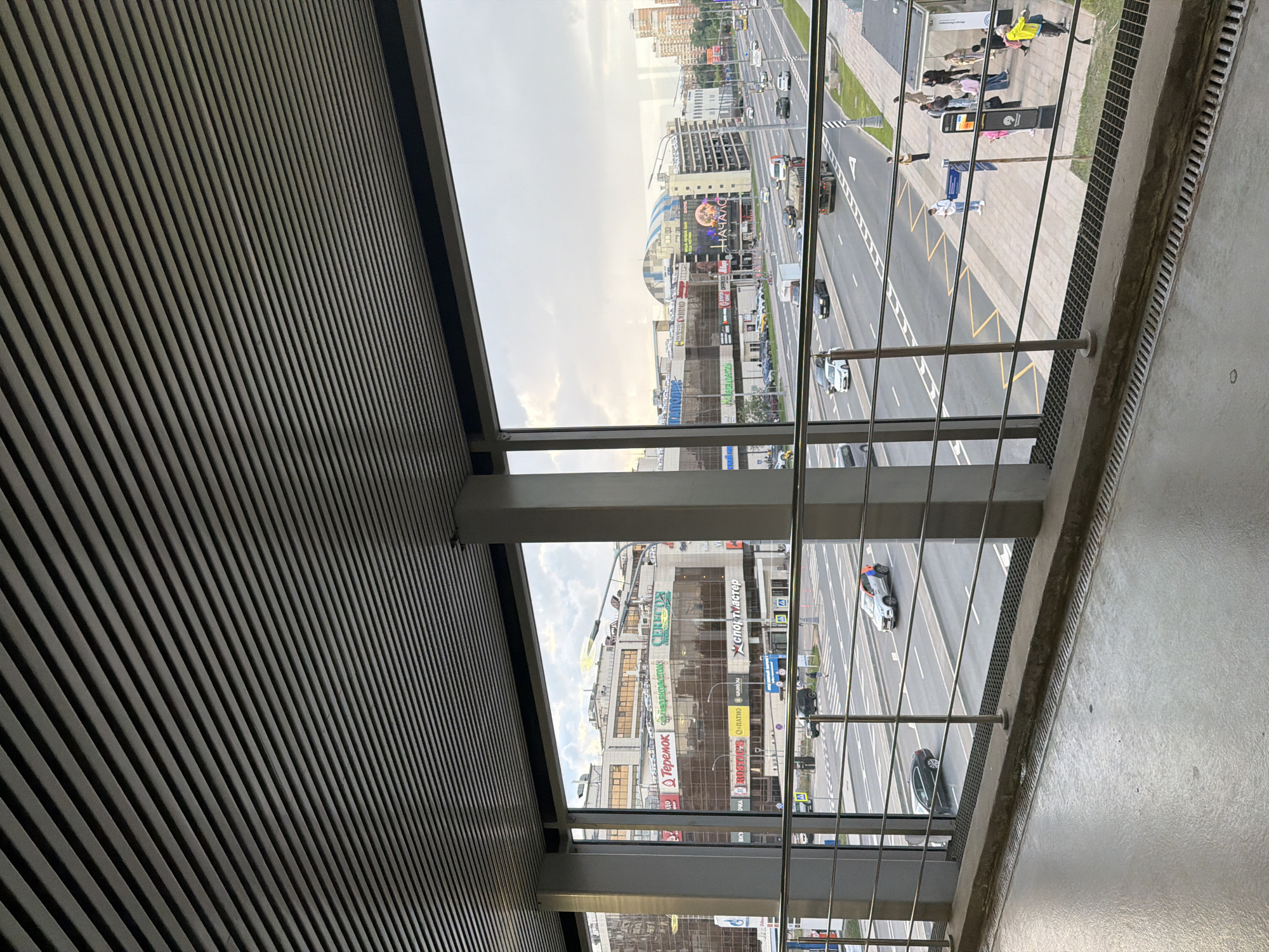 The image is a wide-angle view looking out from a covered walkway or bridge. The foreground features a concrete floor and a metal railing. The background shows a multi-lane road with various vehicles, including cars, a truck, and a bus. On the other side of the road, there are several large, modern buildings with glass facades, suggesting a commercial or urban area. Some of the buildings have visible signage. People are visible on the sidewalk and at a bus stop. The sky is overcast with some clouds. The overall scene depicts a typical urban environment with moderate traffic and pedestrian activity.