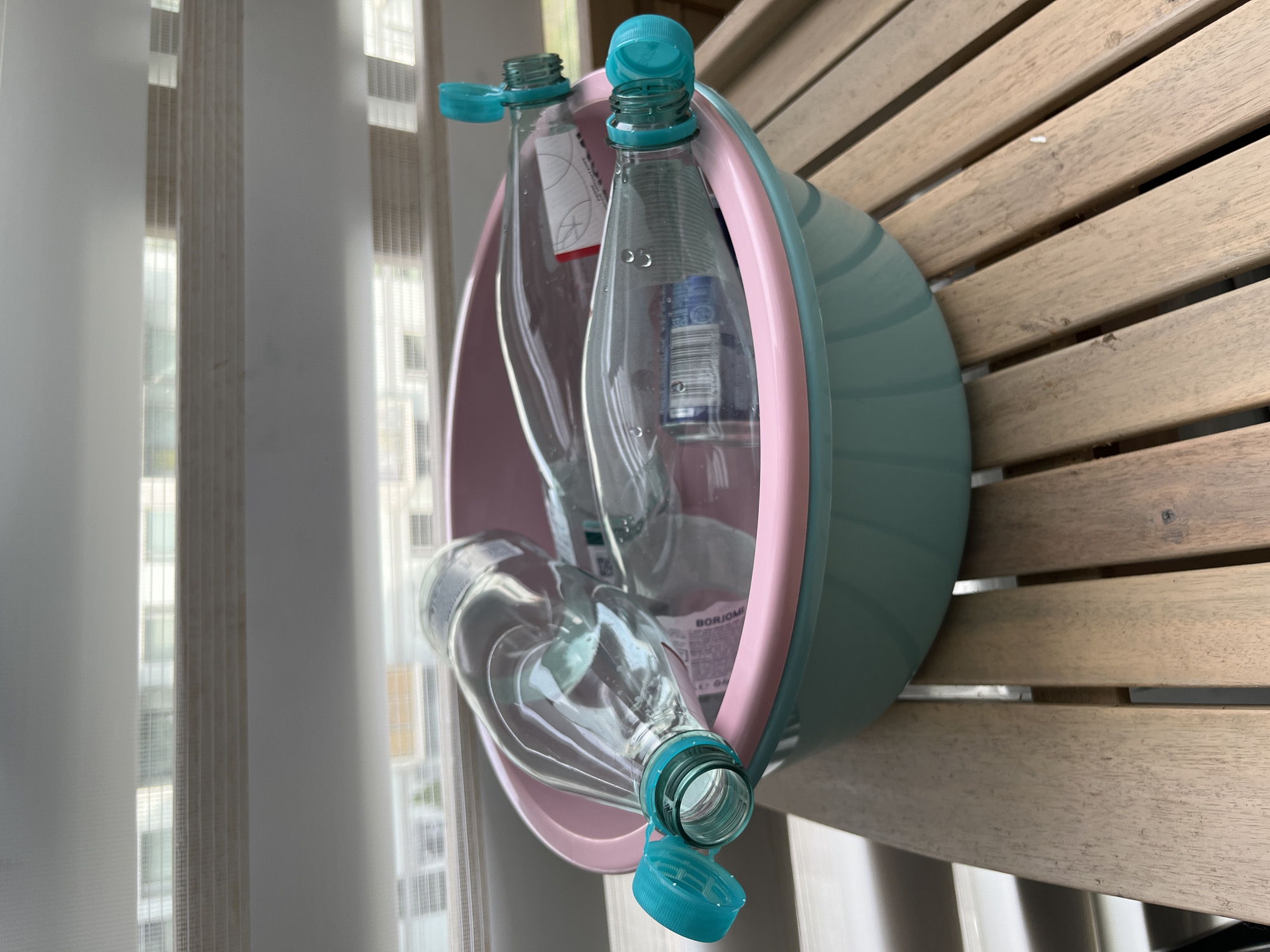 Four plastic water bottles with teal caps are submerged in a pink and teal plastic basin. The basin rests on a wooden slatted surface, possibly a table or deck. The bottles appear to be empty and are lying on their sides, with their caps open or partially open. Water droplets are visible on the surface of the bottles. In the background, a window with horizontal blinds is visible, revealing a blurry view of buildings. The lighting suggests it is daytime. There is no indication of the location being Helsinki, Finland.