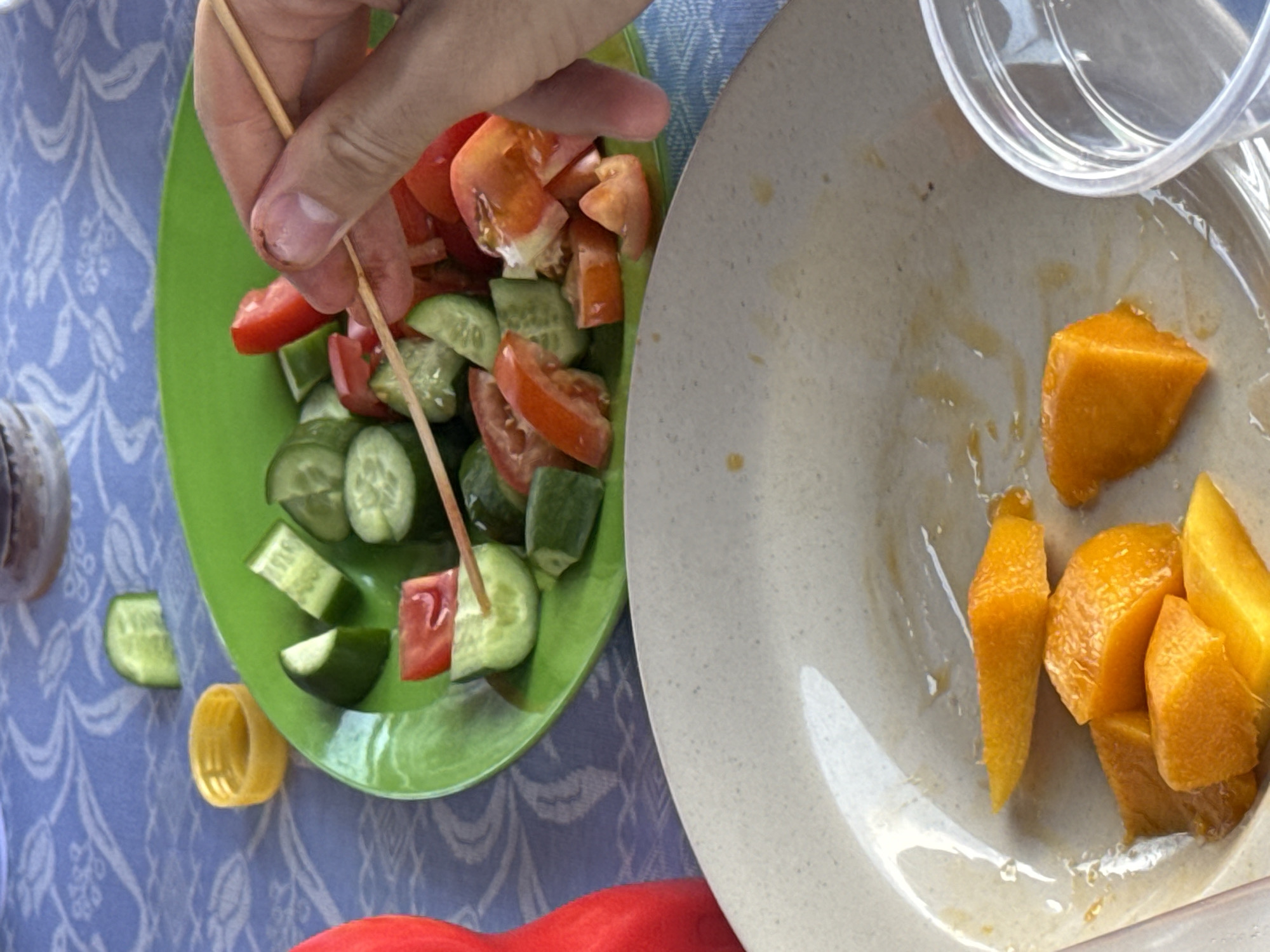 The image depicts a close-up of two plates on a blue patterned surface, possibly a tablecloth, in an undetermined location. A person's hand is visible holding a wooden skewer, piercing a piece of tomato and cucumber from a green plate. The green plate contains chopped tomatoes and cucumbers. The second plate, off-white in color, contains chunks of orange fruit, likely mango. A clear plastic container is partially visible above the fruit plate. A piece of cucumber and a yellow bottle cap rest next to the green plate on the tablecloth. A red object is partially visible at the bottom center of the image. The location of Bac Nha Trang Ward, Vietnam, is not deducible from the visual information within the image.