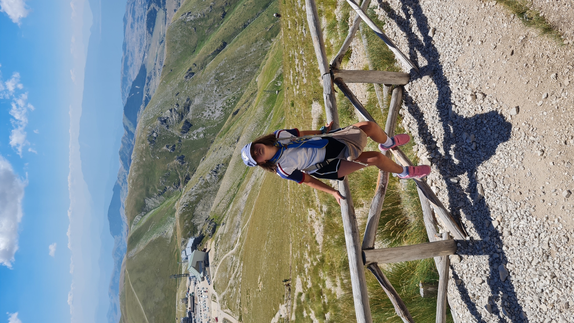 A young girl, wearing a blue baseball cap, a white and blue short-sleeved shirt with red trim, black shorts, and pink shoes, is perched on a wooden railing on a mountainside. She is looking up and to her right, with a light-colored crossbody bag slung over her shoulder. Her legs are bent, with her pink-shoed feet dangling over the railing. The railing is made of rough-hewn wooden posts and rails, with a rocky, gravelly path beneath it. The background reveals a vast mountainous landscape with rolling green hills, dotted with sparse vegetation and rocky outcrops. In the distance, several layers of hazy blue mountains fade into the horizon under a bright blue sky with scattered white clouds. To the left, a modern building complex with a white and gray structure and a parking lot is visible on a lower slope, suggesting it might be a ski resort or a visitor center. The scene appears to be located in the mountainous region surrounding L'Aquila, Italy, possibly at a high-altitude viewpoint.