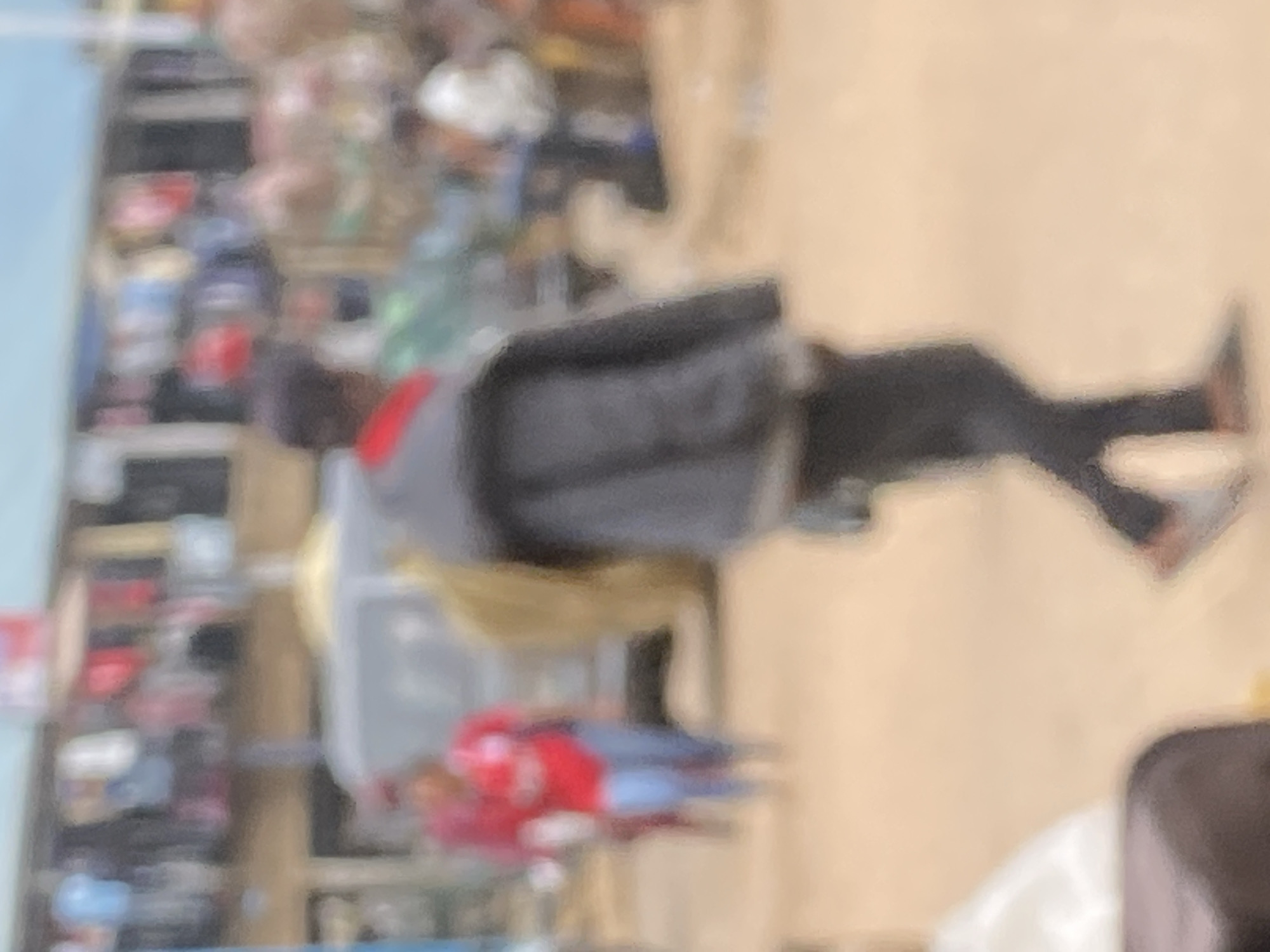 This is a medium shot of an outdoor market scene in Bukuru, Nigeria. The image is out of focus, but it appears to depict a busy commercial area with stalls and people. In the foreground, a person wearing a dark jacket and dark pants is walking away from the camera. Behind them, another person in a red shirt and blue pants is visible. The background shows a row of market stalls with various items displayed, and other people milling about. The ground is a sandy or dirt surface.