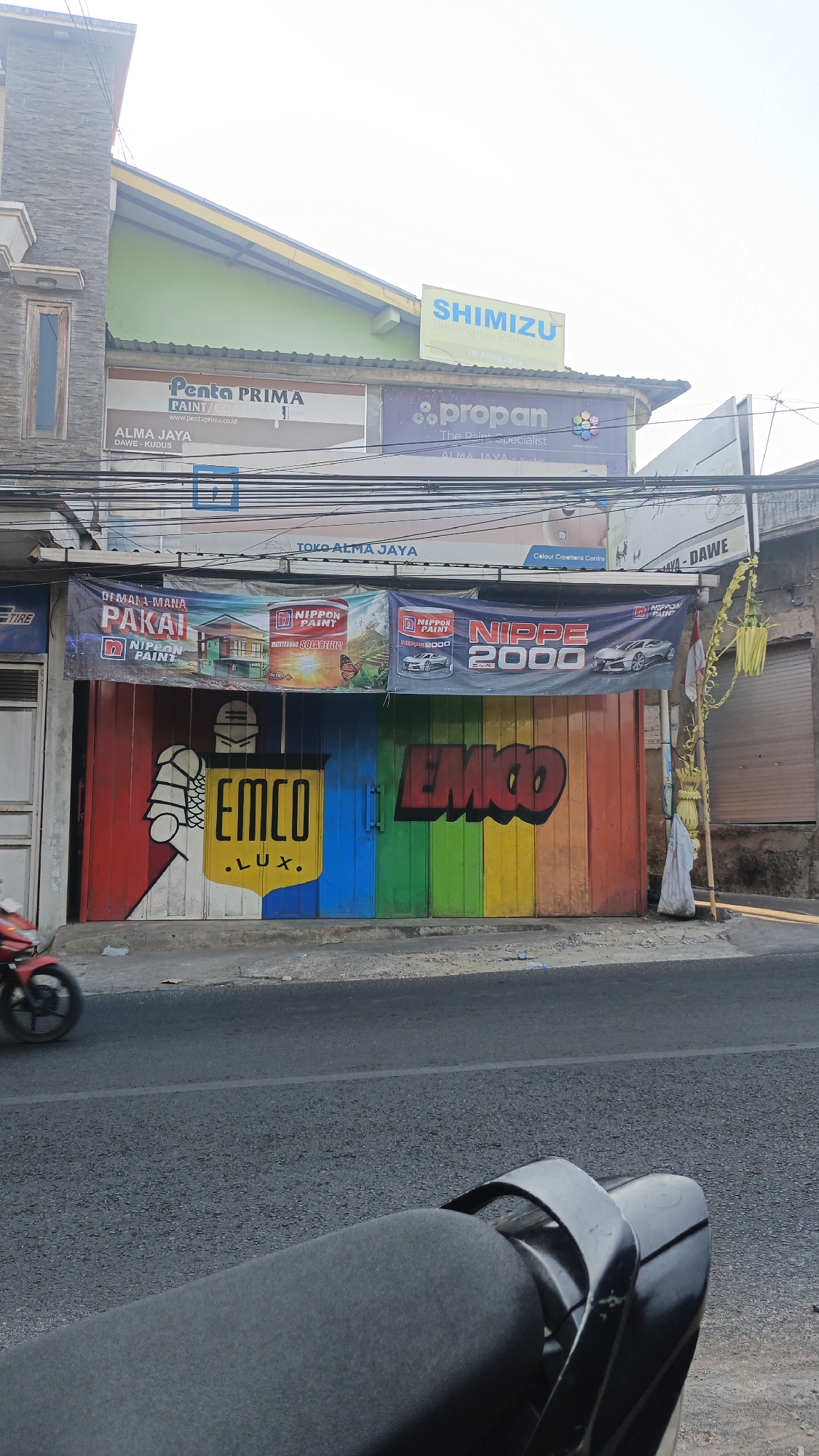 This is a photograph of a street scene in Dawe, Indonesia, featuring a commercial building with a brightly painted roll-up door. The building has several signs advertising paint products, including "Penta PRIMA PAINT," "Shimizu," and "Propan." A banner above the roll-up door displays "Nippon Paint" and "Nippe 2000." The roll-up door itself is painted with large, colorful graphics. On the left side of the door, there's a graphic of a hand holding what appears to be a paintbrush, and the word "EMCO" in large yellow letters with ".LUX." below it. To the right of this, the word "EMCO" is again depicted in red graffiti-style lettering, spanning across several color panels of the door. The street in front of the building shows a red motorcycle in motion on the left side of the frame. The overall environment suggests a commercial area with shops lining the street.