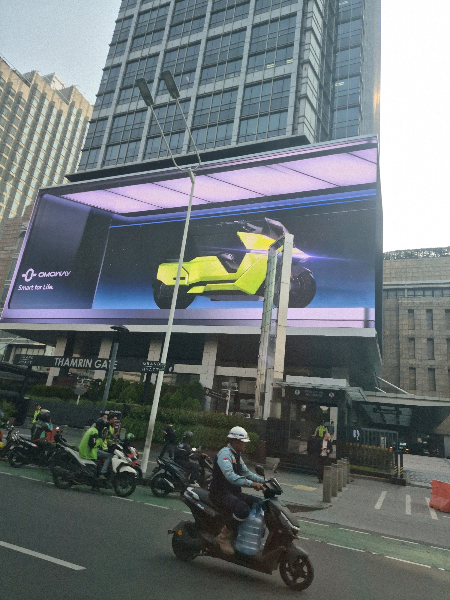 The image depicts a street scene in Central Jakarta, Indonesia, with a prominent digital billboard advertising "OMOWAY Smart for Life." The billboard displays a futuristic, lime-green electric scooter against a dark, starry background. Below the billboard, the signage for "THAMRIN GATE" and "GRAND HYATT" is visible, indicating a location in the bustling Thamrin area.
On the street in front of the buildings, several motorcycles are parked and in motion. One motorcycle rider, wearing a white helmet and a light blue and white uniform, is prominently featured in the foreground, carrying two large water bottles attached to the side of his vehicle. Other individuals on motorcycles, some wearing bright green vests, are also visible, suggesting a busy urban environment with a mix of commuters and delivery services. The architecture of the buildings in the background, with their glass facades and geometric designs, points to a modern business district. The overall impression is one of a dynamic, contemporary urban setting in Indonesia's capital.