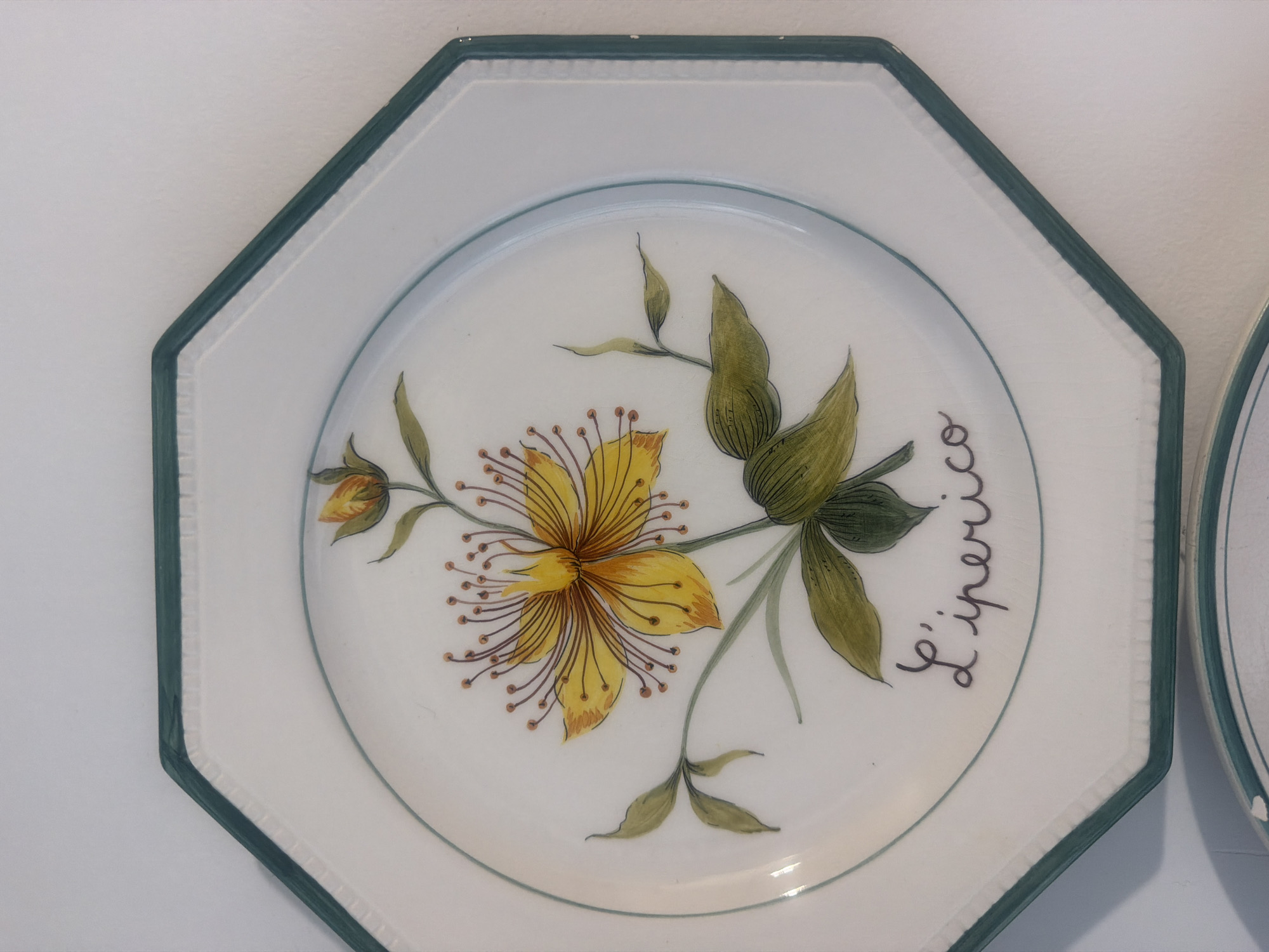 The image shows a close-up, eye-level, medium shot of an octagonal ceramic plate with a green rim. The plate is white and features a hand-painted yellow flower with prominent stamens and orange tips. The flower is depicted with green leaves and stem. Below the flower, written in dark cursive script, are the words "L'iperico". The plate is mounted on a white wall. Another ceramic plate, partially visible at the bottom of the frame, also has a green rim and white surface. The overall setting appears to be indoors. There are no people present in the image, and no specific location within Mandra, Greece, can be identified from the visual information provided.