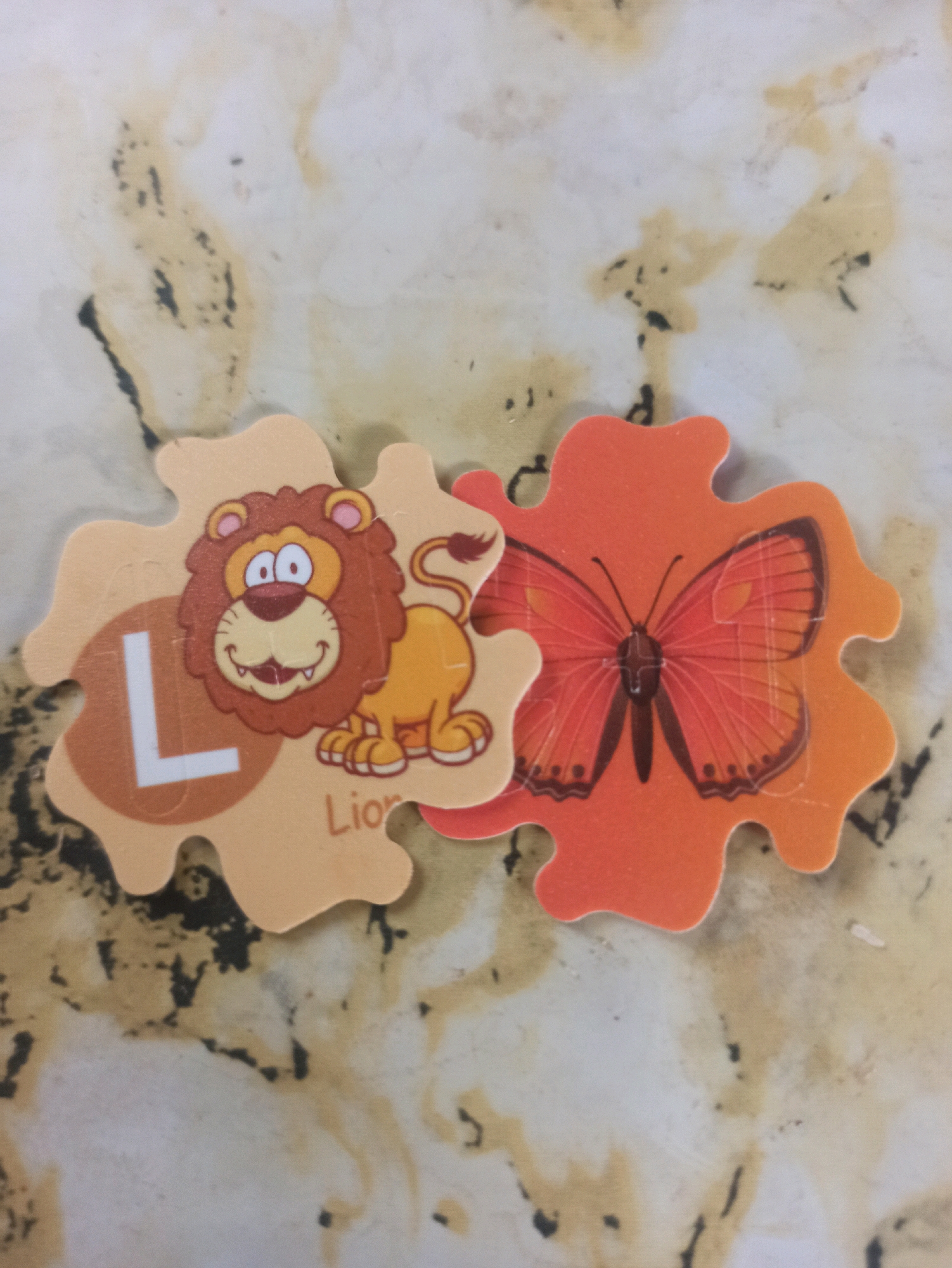 The image shows two puzzle pieces on a marbled surface. The left puzzle piece is light brown and features a cartoon lion with a mane, a smile, and wide eyes. The letter "L" in white is visible on the left side of this piece. Below the lion, the word "Lion" is partially visible. The right puzzle piece is orange and depicts a bright orange butterfly with black markings. The two puzzle pieces are interlocked. The background surface has irregular brown and black markings. The scene is indoors, on a flat surface. There are no people or animals present. The location is not specified in the image.