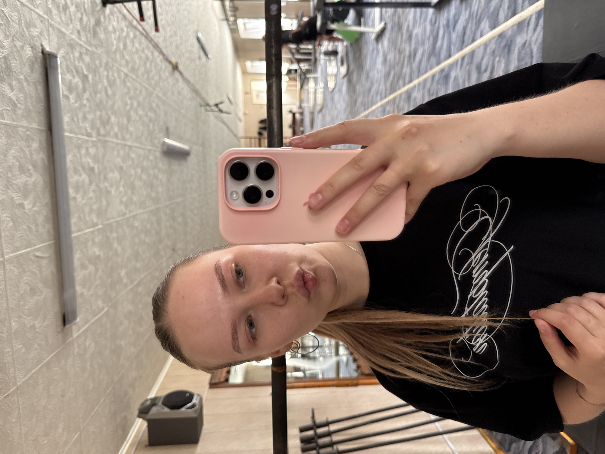 A woman is taking a selfie with a pink-cased iPhone. She has blonde hair and blue eyes. She is wearing a black shirt with white writing on it. The selfie is taken in a gym in Chaikovsky, Russia. Gym equipment is visible in the background, as well as a textured wall with metal bars. There is a grey trash can near the floor.