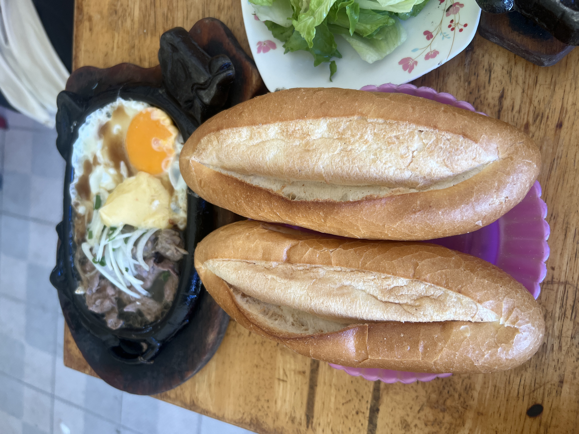The image displays a meal arranged on a light brown wooden table, consistent with a casual dining setting in Nha Trang, Vietnam. The central subject is a black oval cast iron sizzling plate, resting on a wooden trivet, containing sliced cooked beef topped with sliced white onions and green herbs. Adjacent to the beef are two distinct egg preparations: one fully cooked fried egg with a prominent orange yolk and set white, and another lighter yellow mass, possibly a pat of butter or a soft-scrambled egg. Two elongated, golden-brown baguettes, split lengthwise to reveal their white interior, are positioned side-by-side on a purple scalloped plastic tray. To the right, a small white ceramic plate with a pink floral pattern holds fresh green lettuce leaves. The background shows a light-colored tiled floor and blurred white architectural elements, indicating an open-air or semi-enclosed restaurant environment. No human subjects or active interactions are visible.