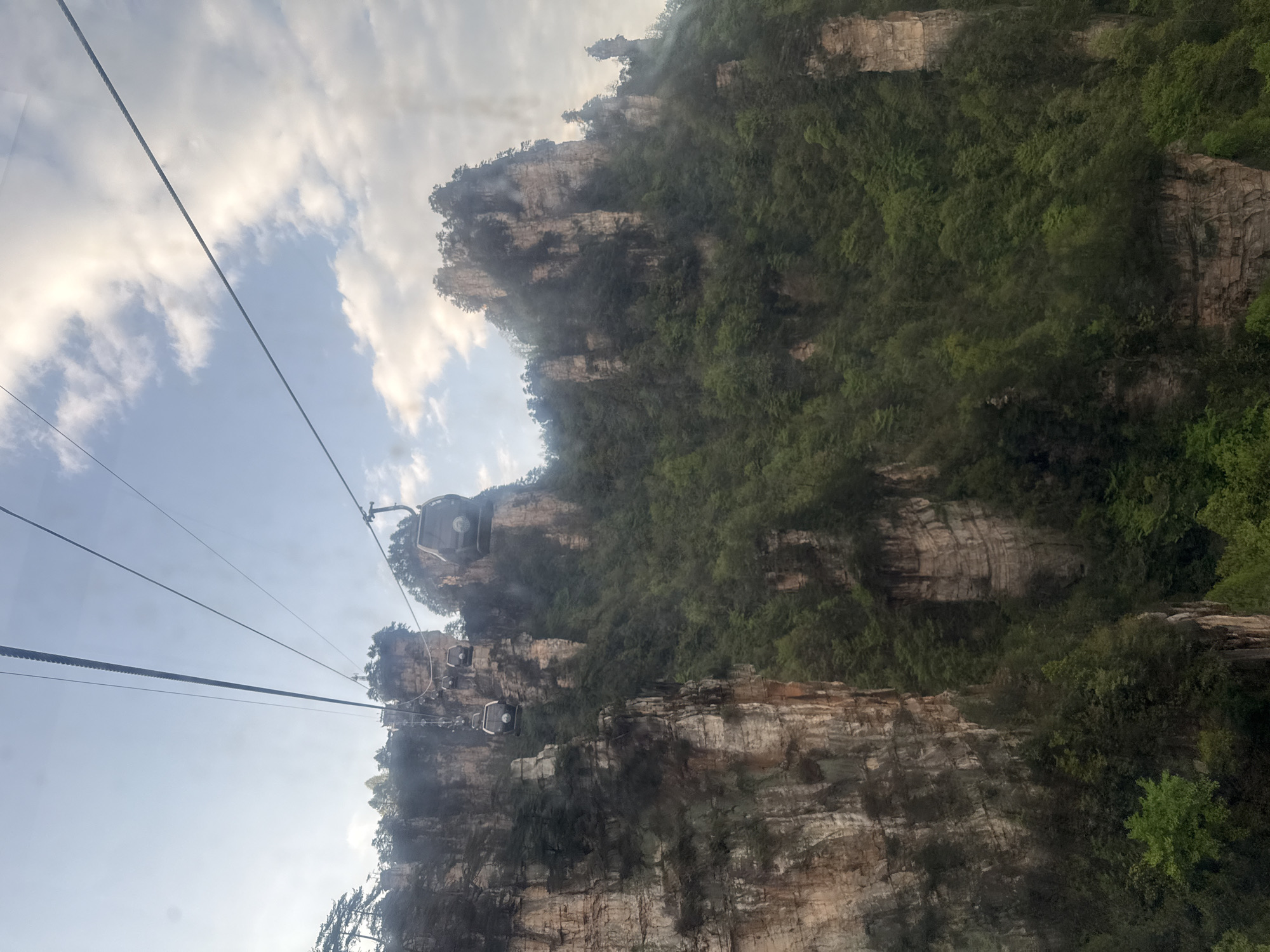 The image captures a scene of the Zhangjiajie National Forest Park in Wulingyuan, China. The photo shows a series of pillar-like rock formations covered with green vegetation. A cable car system with several cars is visible traversing the landscape in the foreground. The sky is visible in the background, partially obscured by clouds.