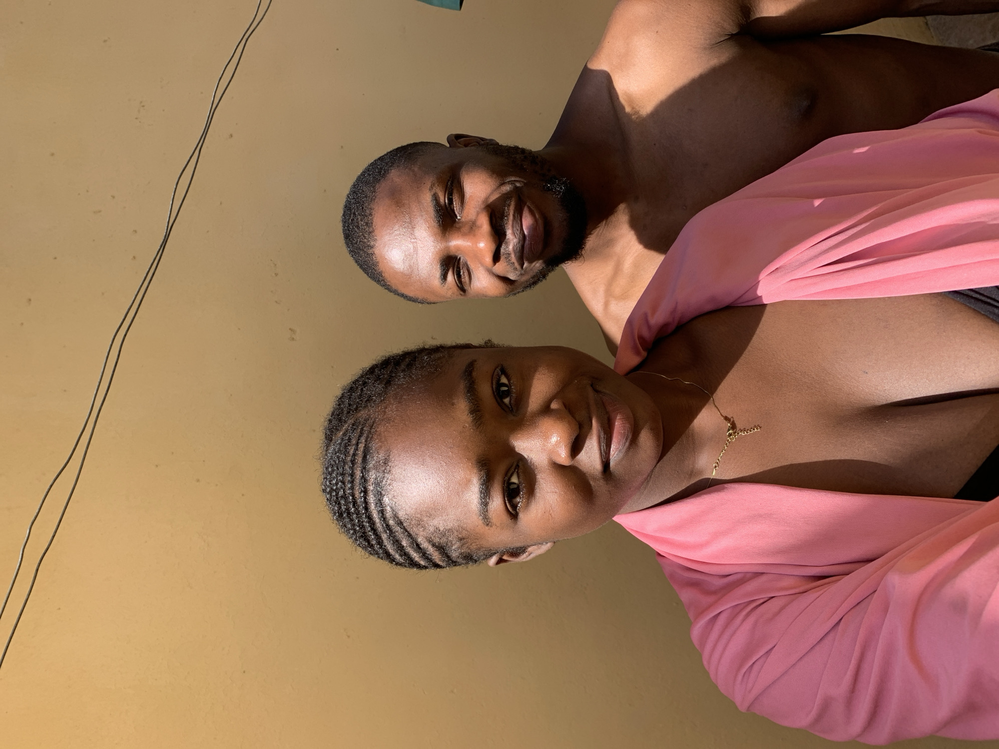 A couple is posing for a photograph in Rafin Par, Nigeria. A woman with braided hair is in the foreground wearing a pink top. A man with a beard stands behind her, shirtless. The background is a yellow wall with a powerline.