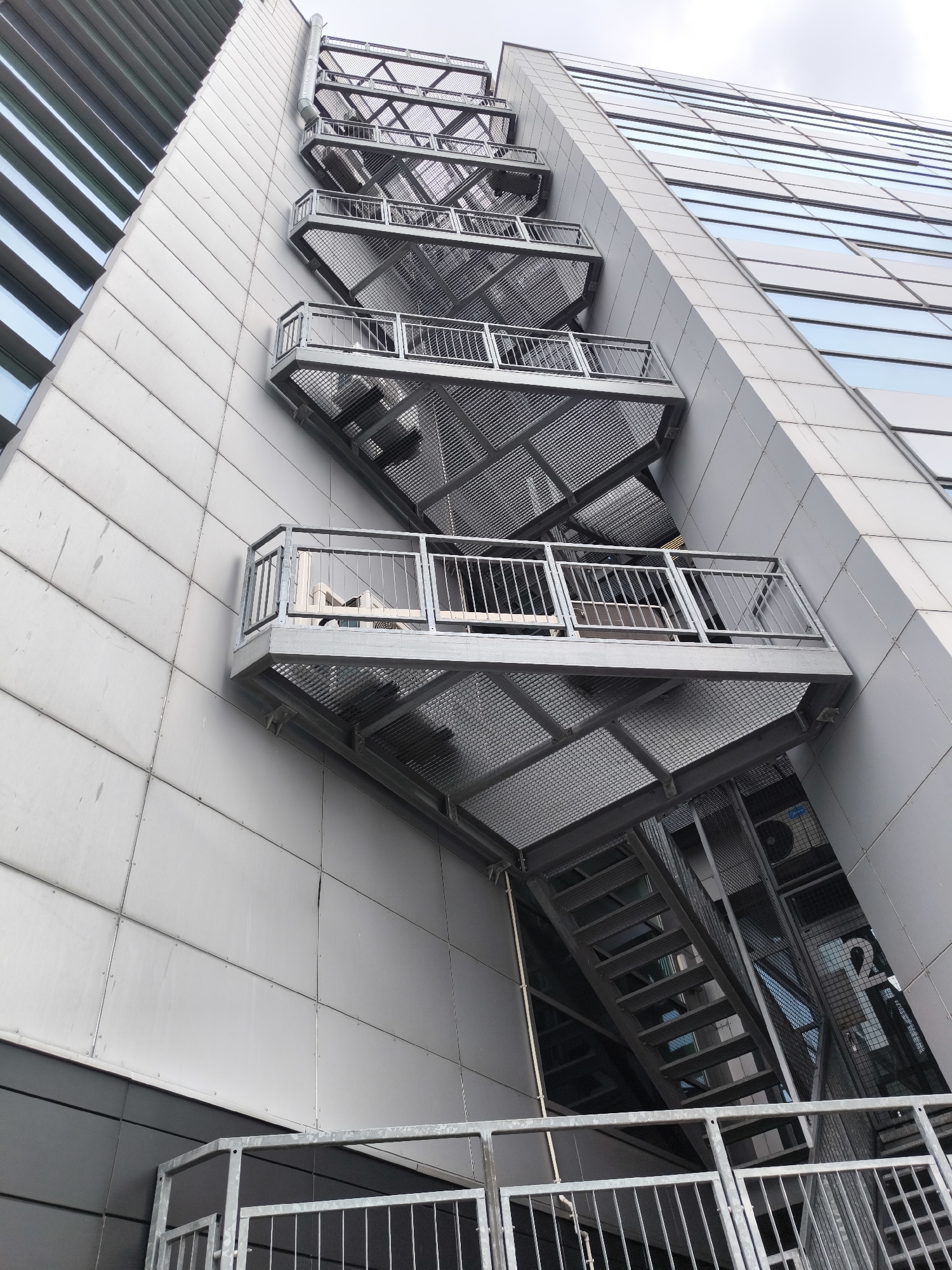 An exterior shot shows a section of a tall building with a metal fire escape attached to one side. The building has a facade of gray rectangular panels. The fire escape consists of multiple platforms with metal railings, connected by metal stairs. The stairs are also metal and have a perforated design. Part of the building next to the fire escape has reflective windows, reflecting the sky. The fire escape extends upwards out of the frame. The building's location is Riga, Latvia.