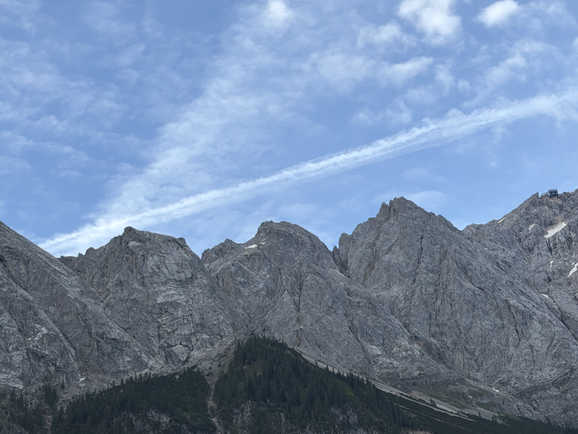 The image shows a wide shot of a mountain range under a bright blue sky with scattered white clouds and a faint contrail. The mountains are rugged and rocky, with steep slopes and jagged peaks. Dark green coniferous trees cover the lower slopes of the mountains. In the upper right portion of the image, a small building with a dark roof is visible on the summit of one of the peaks. This appears to be a view of the Zugspitze, Germany's highest peak, from Grainau, Germany. There are no people or animals visible in the image.