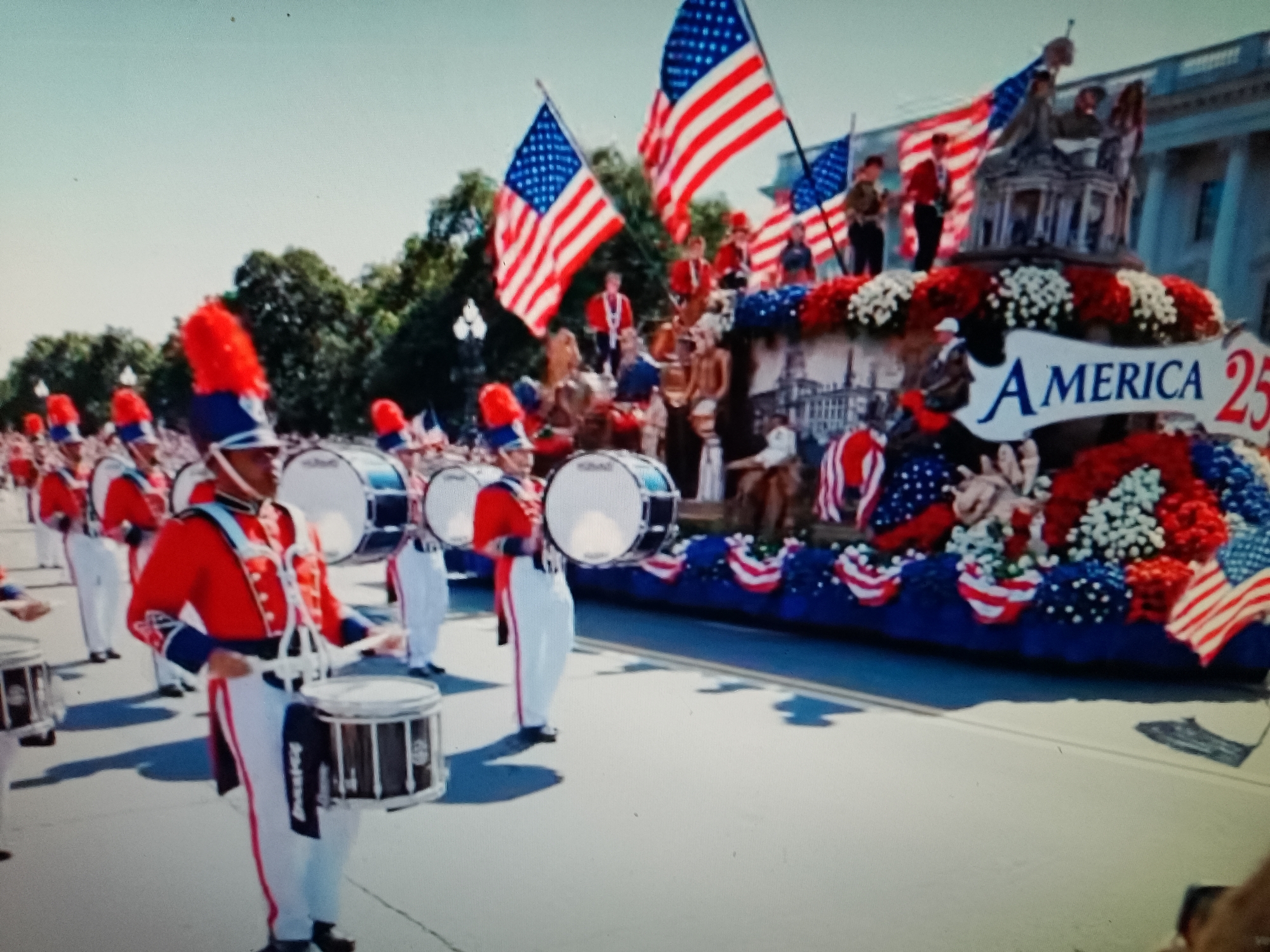 The image depicts a parade in progress, likely a patriotic celebration given the prominent display of American flags and red, white, and blue decorations. A marching band, composed of individuals in vibrant red and blue uniforms with tall, feathered hats, is visible in the foreground, several members playing drums. Behind the band, a decorated float is adorned with flowers and American flags, featuring a large banner that reads "AMERICA 25". People are gathered on and around the float, and spectators appear to be present in the background, though their details are less clear. The overall scene suggests an outdoor event under a bright, possibly sunny sky, with trees visible in the distance. The mention of Chiajna, Romania, is not supported by any visual elements within the provided image.
