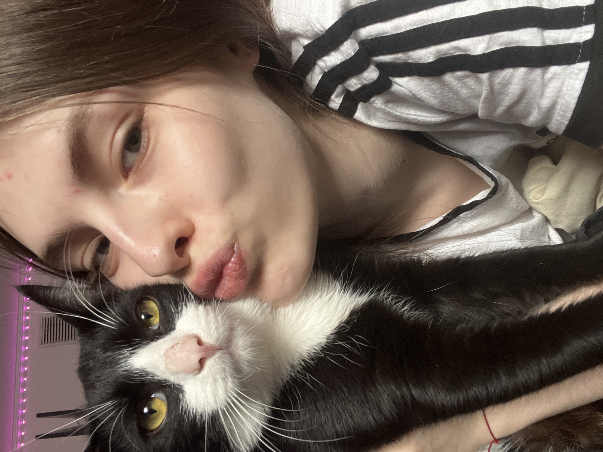The image is a close-up, rotated selfie featuring a young person and a black and white tuxedo cat in an affectionate pose. The person, who has fair skin and light brown hair, is lying down with their head tilted, making a "kissing" face with pursed lips towards the camera. They are wearing a white t-shirt with black horizontal stripes, and a small red string or bracelet is visible on their wrist.
The cat is positioned closely beside the person's face, also looking towards the camera. It is a tuxedo cat with mostly black fur, a white chest, a white blaze on its nose, and prominent white whiskers. Its eyes are a striking bright yellow. Both the person and the cat appear calm and relaxed, conveying a strong sense of affection between them.
The setting is an indoor, softly lit environment, likely a bed or couch, suggested by the white and light-colored fabrics in the background. A blurred strip of pinkish-purple light, possibly from LED strip lights, is visible on the far left edge of the frame. No discernible text is visible in the image. This scene takes place in Saint Petersburg, Russia.