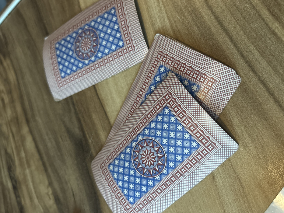 Three distinct groupings of playing cards are displayed face-down on a light-brown wooden surface in Pervomaiskoe, Russia. Two stacks of cards are positioned in the foreground, with one partially overlapping the other towards the lower-left of the frame. A third, smaller stack or single card is visible further back, towards the upper-right. All visible card backs feature an identical design: a central circular medallion with a multi-pointed star motif rendered in red, grey, and light blue, set against a dark blue background. This central element is encompassed by a field of light blue diamond patterns on a dark blue background, bordered by an inner strip of repeating red squares and an outer fine cross-hatched red border. The cards show signs of wear along their edges and corners, and a noticeable brown discoloration is present on the bottom right corner of the foremost foreground card. The wooden surface exhibits a distinct grain pattern, running predominantly horizontally across the frame. No individuals or active interactions are visible.