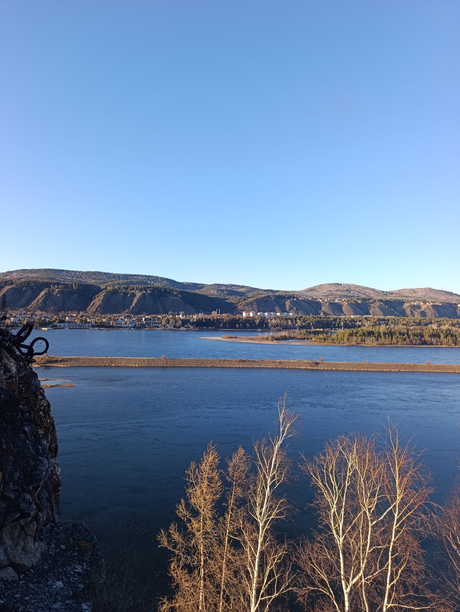 A panoramic view of the Yenisey River and the Krasnoyarsk skyline in Russia. Clear blue sky dominates the scene. The river is the primary element, spanning much of the image width. A long, thin stretch of land divides the river into two channels in the middle ground. In the background, there is a line of mountains with urban development on its slopes. In the foreground, trees without leaves and part of a rocky cliff are visible.