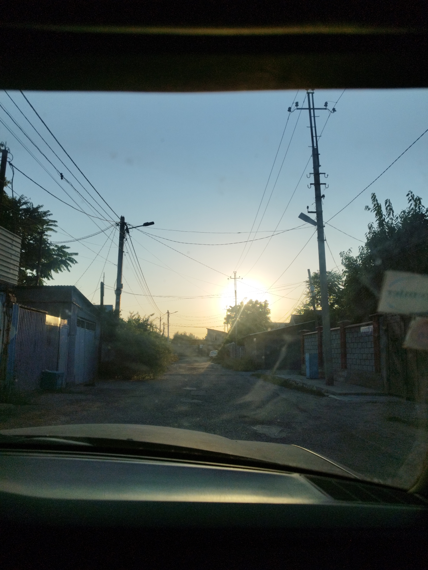The image shows a street scene in the city of Shymkent, Kazakhstan. The sun is setting, casting a warm glow over the street and surrounding buildings. The street is unpaved and appears to be in disrepair, with potholes and cracks visible. There are several power lines crisscrossing the sky, supported by wooden and concrete poles. On either side of the street are buildings of various sizes and materials, including corrugated metal structures and brick houses. Some of the buildings appear to be residential, while others may be commercial or industrial. There are trees and overgrown vegetation along the sides of the street, adding a touch of nature to the urban landscape. In the distance, a white car is visible parked on the side of the road. The overall impression is one of a quiet, somewhat neglected street in a residential area of Shymkent.