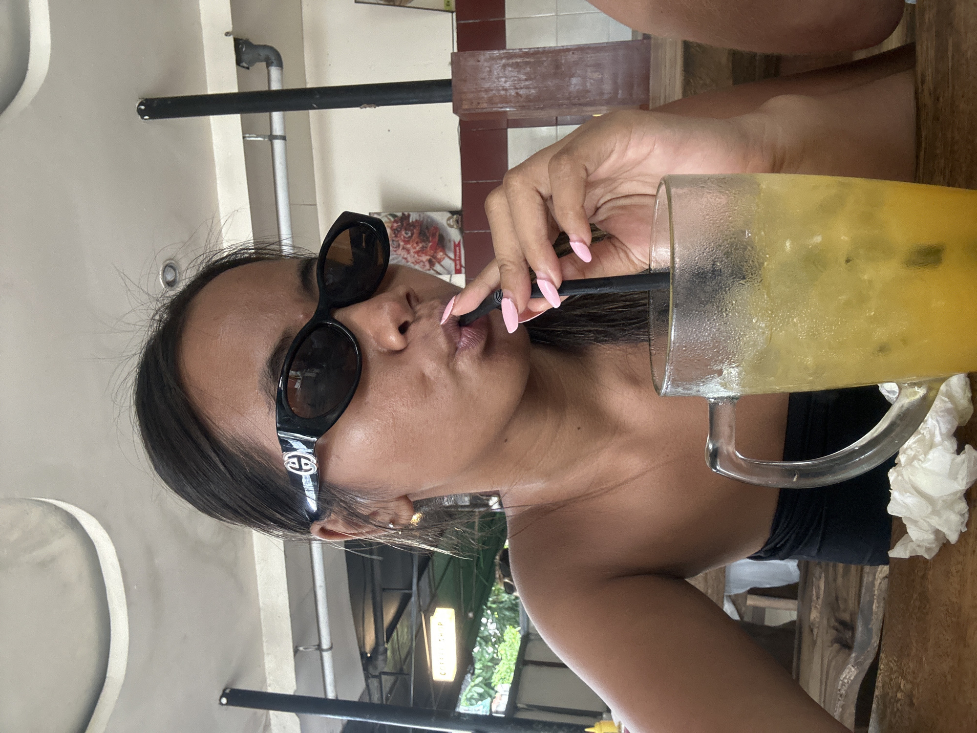 A woman is sitting at a wooden table in a restaurant, wearing black sunglasses and a black strapless top. She is holding a straw with her right hand, with long, pale pink painted nails, and drinking from a tall glass of a yellow, ice-filled beverage. She has dark hair pulled back and is wearing small gold hoop earrings. The background shows a light-colored ceiling, a black pole, and a wall with a framed picture of food. A crumpled white napkin is on the table. The lighting suggests it is daytime.