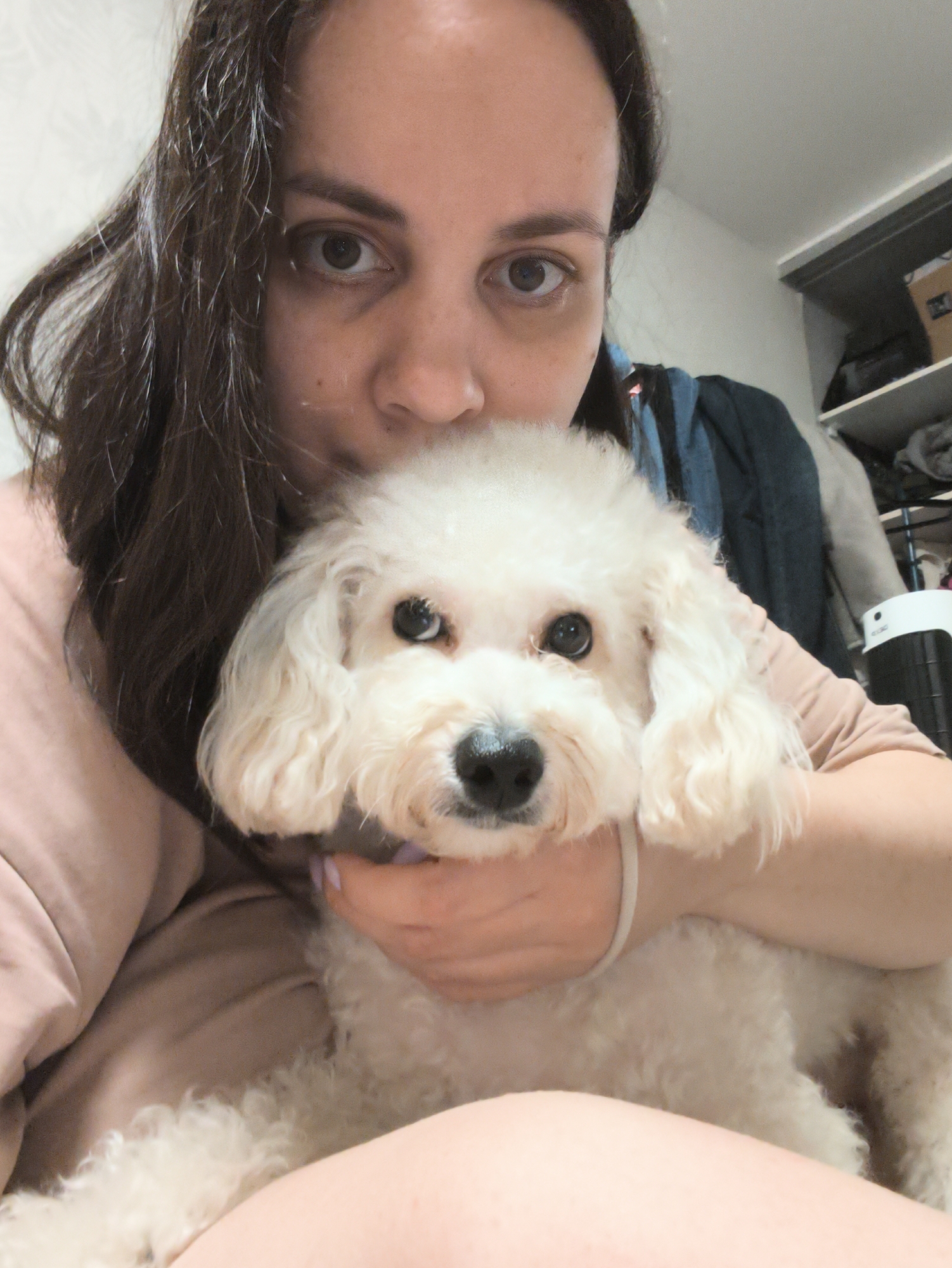 A woman with dark hair holds a small, white, curly-haired dog close to her face. She looks directly at the camera with wide eyes. Her hand supports the dog under its chin. She wears a peach-colored shirt. The dog has a black nose and dark eyes. The background shows a wardrobe or closet with clothes and boxes. The environment looks like an apartment or home. There is no indication that the image was taken in the city of Chaikovsky, Russia.