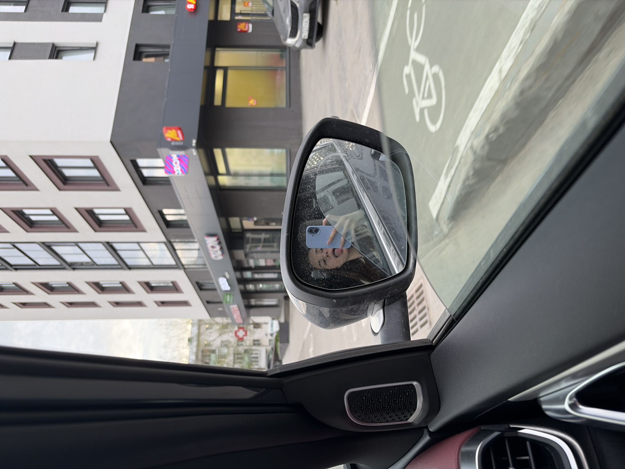 The image is taken from inside a car, looking out the passenger side window. The side mirror reflects a person holding a blue smartphone taking a selfie. Outside, there is a multi-story building with a modern design, a mix of white and dark grey panels, and multiple windows. Signs on the building include the name "OZON." There is a parking area and a bike lane visible on the street. Based on the OZON sign, the image is likely taken in Izhevsk, Russia.