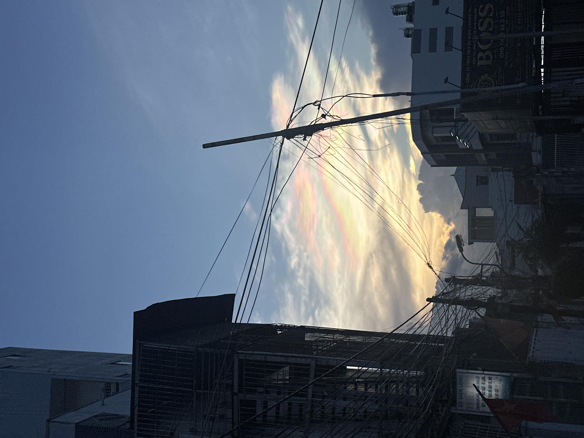 The image shows a street scene in Bac Nha Trang Ward, Vietnam. Overheads are densely filled with electrical wires, obscuring the view of iridescent cloud formations in the sky. A sign for "BOSS Luxury" is visible on a building in the background. A Vietnamese flag is positioned near another building obscured by electrical cables. Other buildings surround the scene.