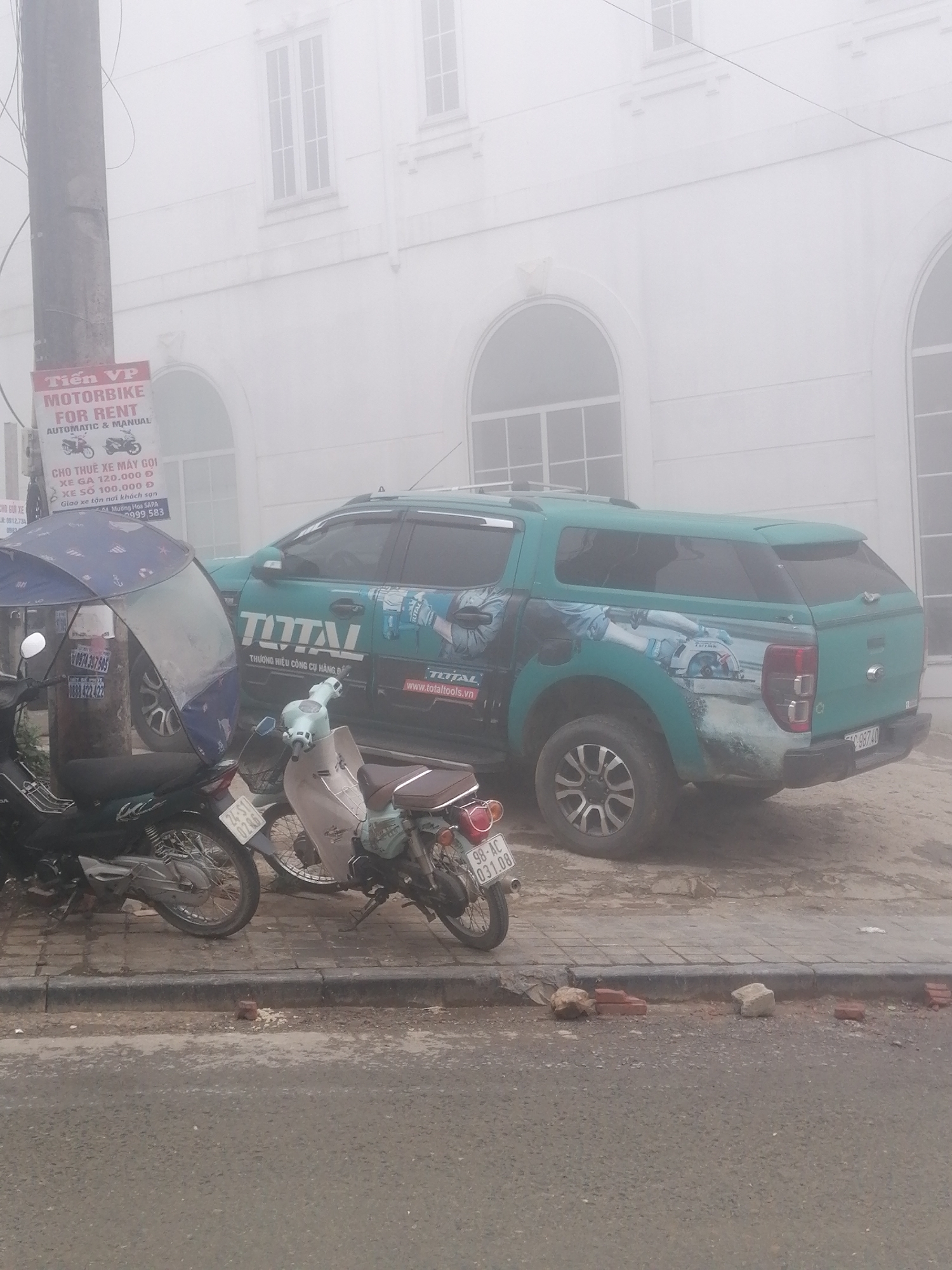 The image captures a misty street scene in Sa Pa Ward, Vietnam, showcasing parked vehicles against a blurred background. The heavy fog diffuses light, giving the scene a soft, almost ethereal quality, and obscures distant details.
In the foreground, two motorbikes are parked on a cracked sidewalk next to a curb. The motorbike on the left is dark-colored and has a blue and white patterned umbrella attached to its handlebars. Next to it is a lighter-colored scooter. Both motorbikes display Vietnamese license plates: "24-S1 0286" on the darker bike and "98-AC 031.08" on the scooter. The sidewalk is in disrepair, with bricks and concrete rubble visible along the curb.
Behind the motorbikes, a teal-colored pickup truck, identified as a Ford Ranger, is parked. Its side is covered with branding for "TOTAL," featuring the text "THƯƠNG HIỆU CÔNG CỤ HÀNG ĐẦU" (Leading tool brand) and "www.totaltools.vn," along with an illustration of a power tool. The truck has a matching teal bed cover.
To the left, a dark utility pole stands, partially obscuring a red and white sign. The sign, written in both English and Vietnamese, advertises "MOTORBIKE FOR RENT AUTOMATIC & MANUAL." Additional Vietnamese text provides prices: "XE GA 120.000 Đ" (Scooter 120,000 VND), "XE SỐ 100.000 Đ" (Geared bike 100,000 VND), and specifies "Giao xe tận nơi khách sạn Mường Hoa SAPA" (Deliver bike to hotel Mường Hoa SAPA), along with several phone numbers including "0974.347.505" and "0999 583."
The background features a large, light-colored building with multiple arched and rectangular windows, fading into the dense fog. No people are visible, and no specific activity is taking place beyond the vehicles being parked. The scene evokes a quiet, possibly early morning or late afternoon, in a foggy urban environment.