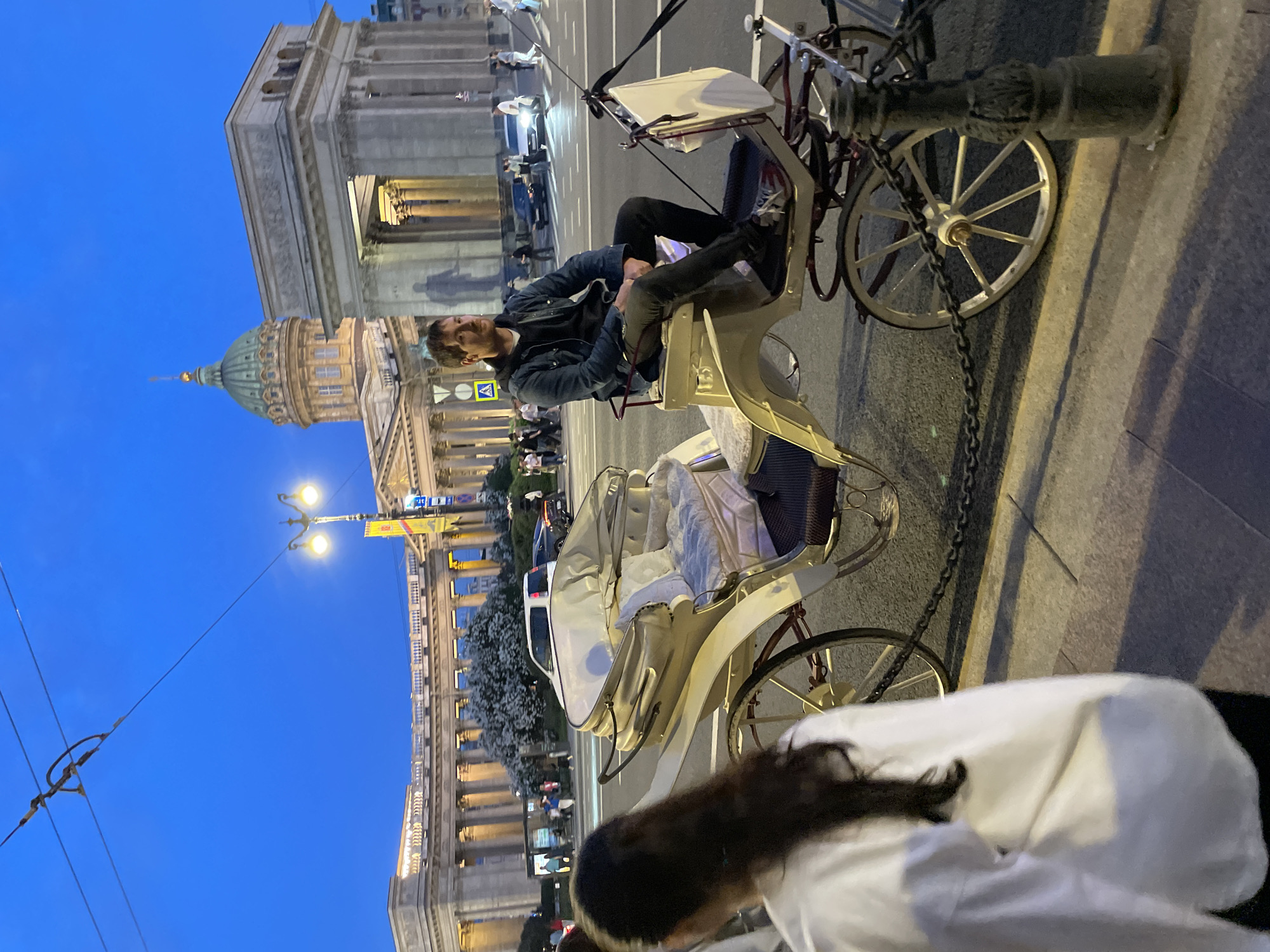 The image depicts an evening or early morning scene in Saint Petersburg, Russia, centered on a street with the Kazan Cathedral and the General Staff Building visible in the background. In the midground, a light-colored, ornate two-seater horse-drawn carriage is parked on the pavement. A man with short brown hair and a mustache, wearing a dark hoodie, a denim jacket, dark trousers, and light-colored sneakers, sits in the front seat of the carriage, looking towards the left. The carriage is secured to a dark bollard in the foreground by a black chain. In the immediate foreground, the back and left shoulder of a woman with long dark hair, wearing a white shirt and dark pants, are partially visible, obscuring the lower left corner of the frame.
To the left of the man and carriage, the distinctive green dome and classical colonnade of the Kazan Cathedral are prominent. To the right, the monumental archway of the General Staff Building (part of the State Hermitage Museum complex) is clearly visible. The sky is a deep twilight blue, indicating dusk or dawn. Streetlights and building lights are illuminated, casting a warm glow on the architectural elements and the street. Numerous pedestrians are visible on the sidewalks and crosswalks in the distance, along with a few cars, including a white van and a dark sedan, on the asphalt road. White flowering trees are situated between the carriage and the Kazan Cathedral's colonnade. Overhead electrical wires are visible against the blue sky.