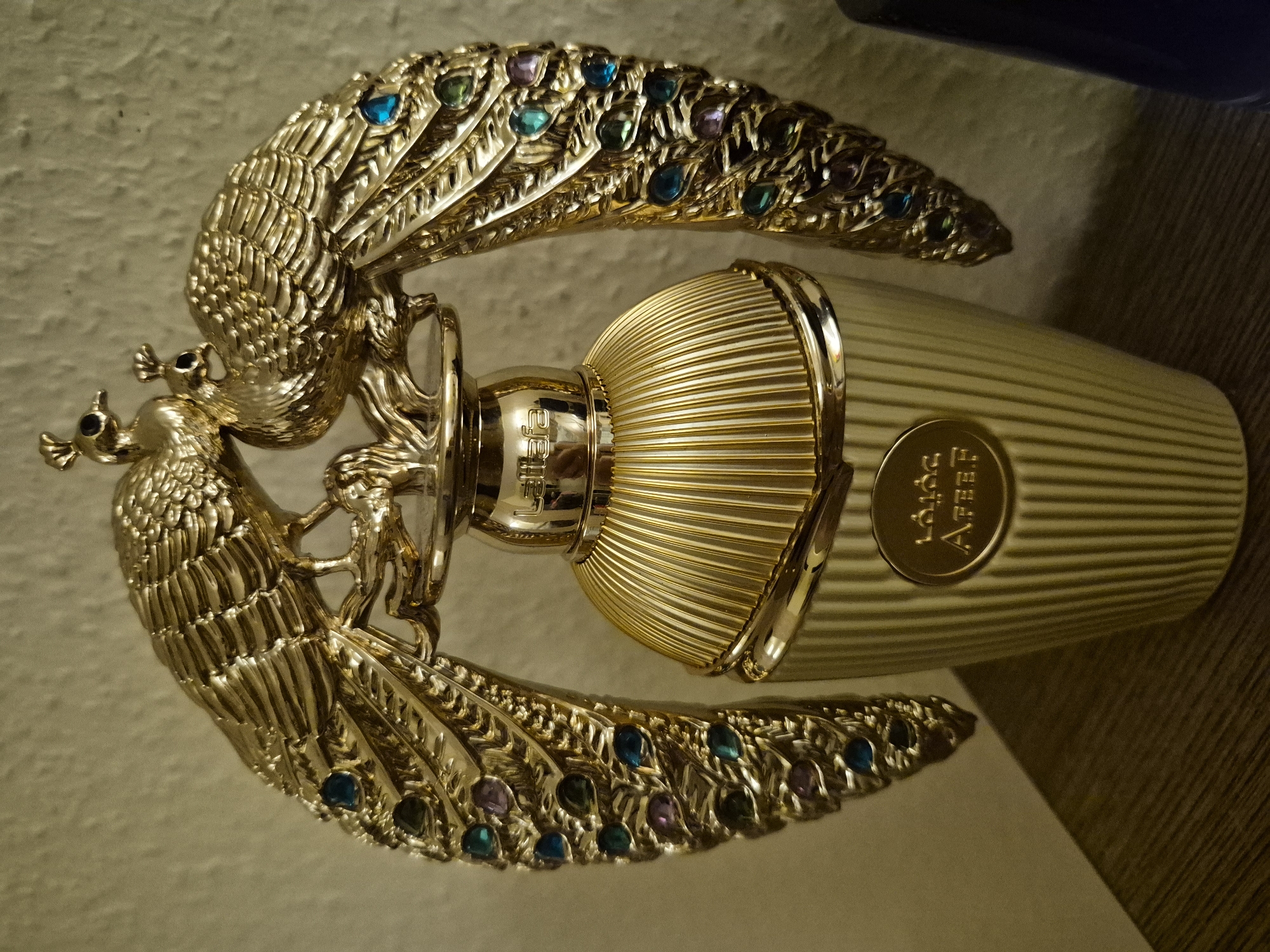An ornate, gold-colored perfume bottle with a peacock-themed design sits on a wooden surface. The bottle's cap is shaped like two intertwined peacocks with jeweled tail feathers. The bottle itself is ridged and has a circular emblem on its front with Arabic writing. The background is a textured, beige wall.