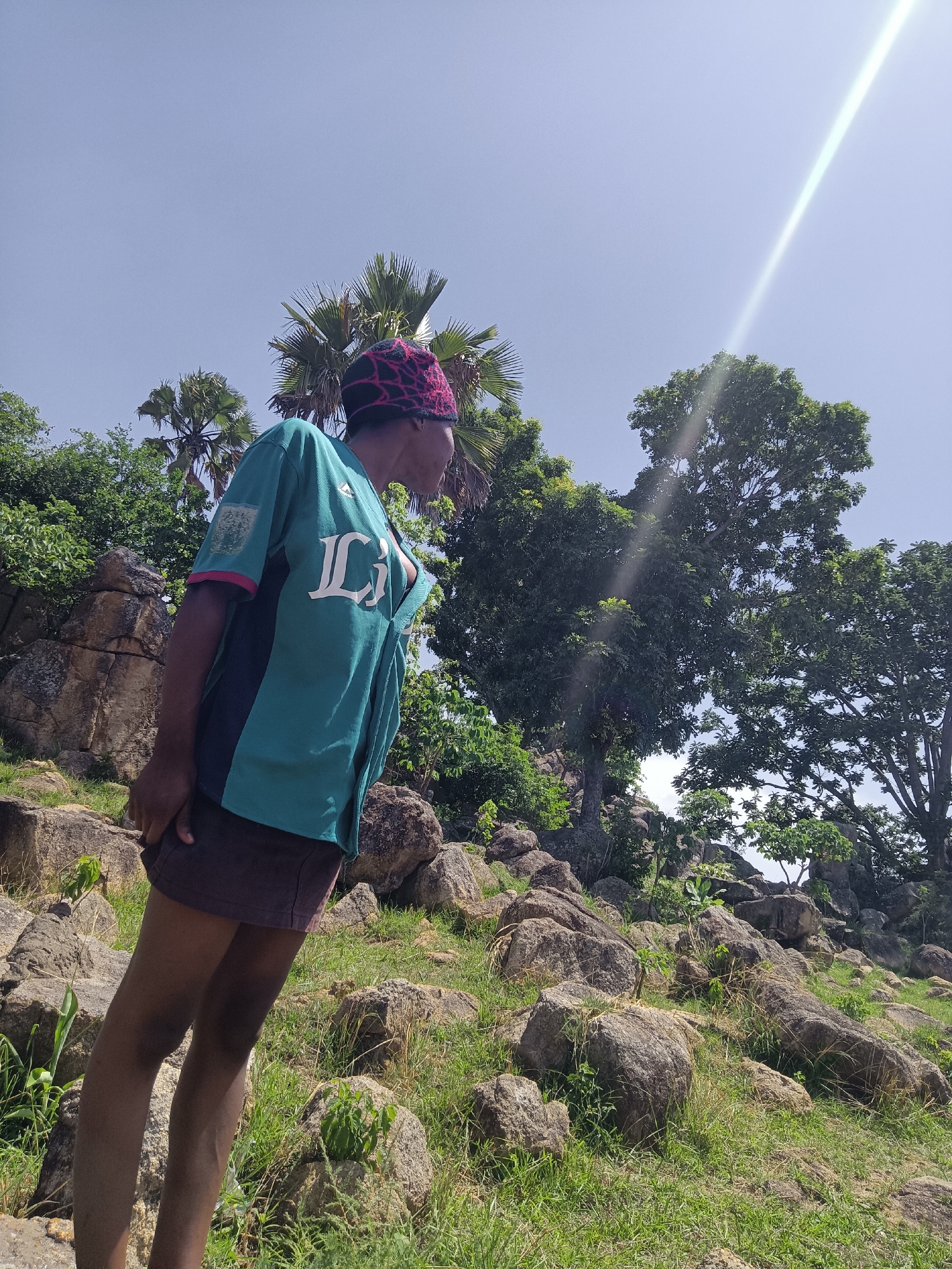 A person stands on a rocky hillside in Du, Nigeria. The individual wears a colorful head covering, a green short-sleeved shirt with a large white "L" on the front, and a dark skirt. The person is looking off to the side, seemingly observing something in the distance. The background features a landscape with scattered trees, rocks, and vegetation under a clear sky with a bright sun streak.