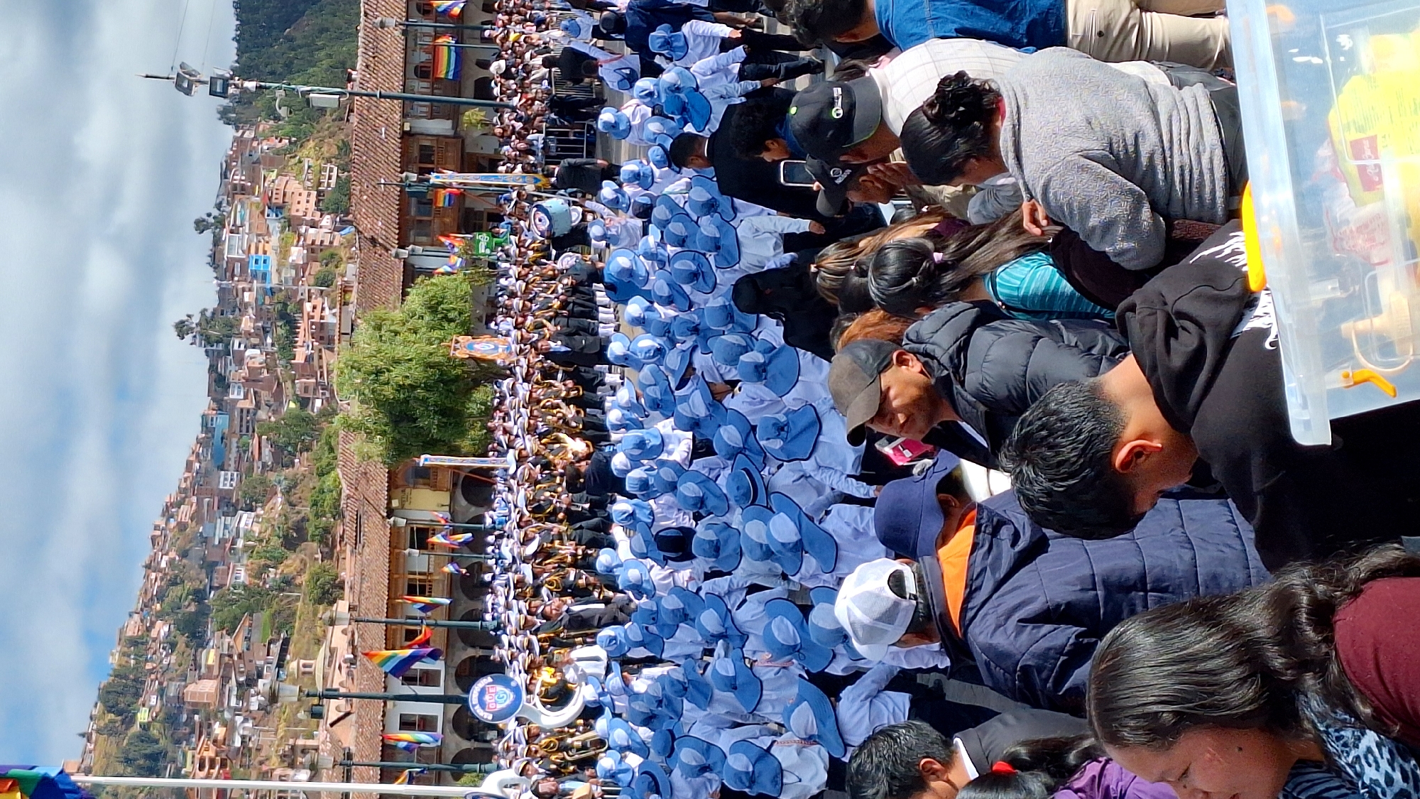 In Cuzco, Peru, a parade is underway in a plaza, with a large crowd gathered to watch. In the foreground, spectators are looking towards the parade. Behind them, a large group of participants in matching blue hats and white uniforms are marching. Further in the background, a brass band in yellow and black uniforms is performing, and beyond that, more participants and spectators are visible. The backdrop of the scene features buildings with traditional terracotta roofs climbing up a hillside, characteristic of Cuzco's urban landscape. The sky is partly cloudy.