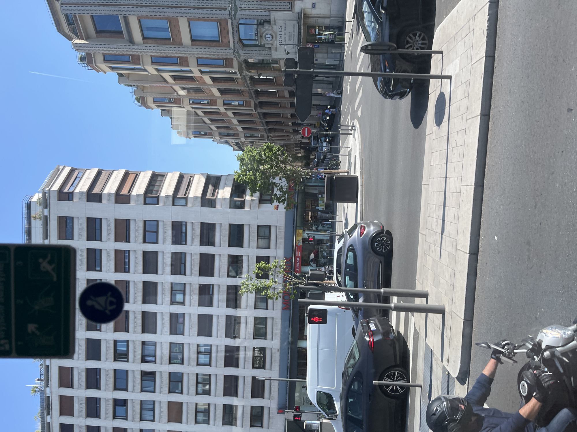 The image depicts a street scene in Neuilly-sur-Seine, France. On the left, a large, light-colored apartment building with numerous windows and brown shutters dominates the background. To the right, a multi-story building with a more ornate, classical facade, featuring brickwork and arched windows, stands. A white van is visible in the middle ground, with traffic lights and pedestrian signals. Several cars are on the road, including a dark sedan, a gray hatchback, and a black sedan. In the foreground, a person wearing a dark blue jacket and a black helmet is riding a motorcycle. The sky is clear blue with a faint contrail from an airplane. The street is paved with asphalt, and there are sidewalks with tiled patterns. Street signs and lampposts are also present. The scene appears to be a sunny day in an urban environment.