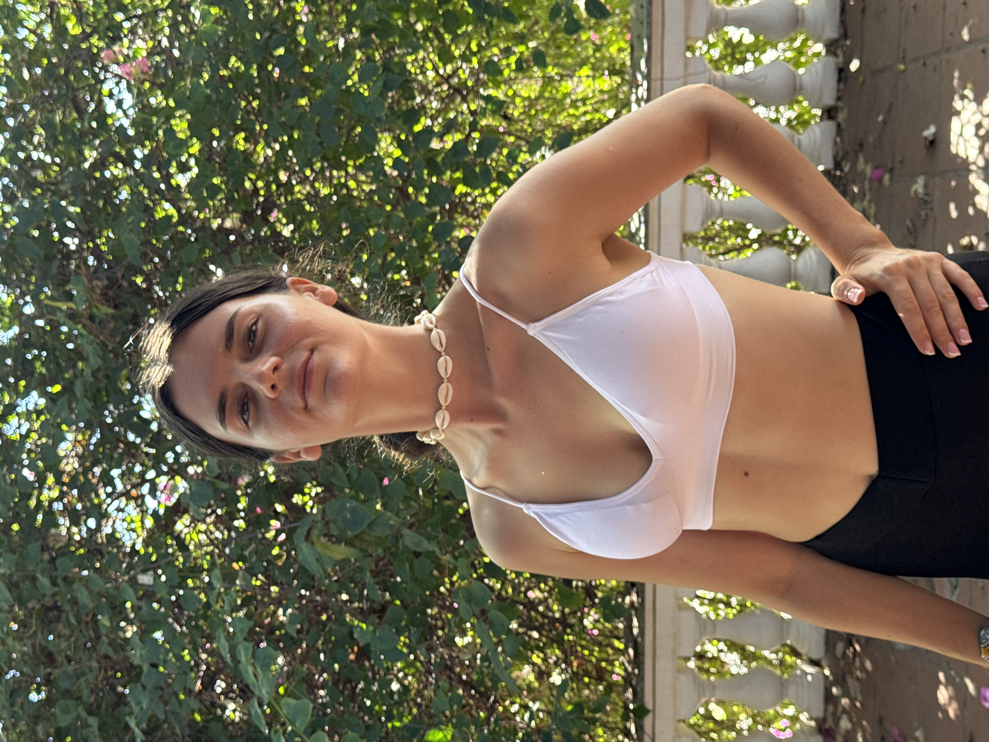 A young woman stands outside in Mũi Né, Vietnam. She is wearing a white top and dark pants. A necklace made of seashells adorns her neck. She is positioned in front of greenery and a decorative white wall. Her right hand rests on her hip. The sunlight filters through the leaves creating dappled shadows.
